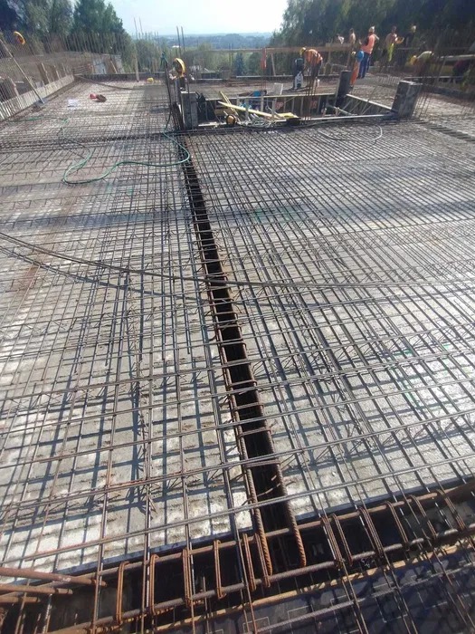 Siatka zbrojeniowa na betonowej powierzchni, widok z góry, z pracownikami w tle. Elementy konstrukcyjne w trakcie budowy.