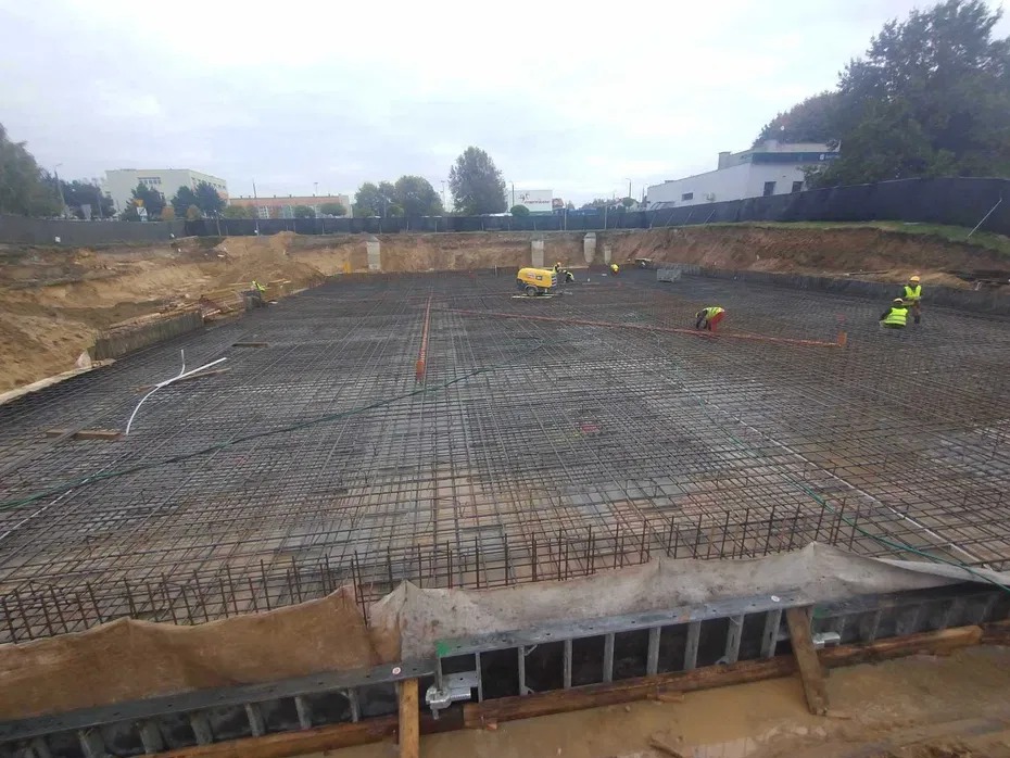 Zbrojenie fundamentów pod budowę w Chorzowie. Widoczna siatka zbrojeniowa, pracownicy w odblaskowych kamizelkach i maszyny budowlane. Teren przygotowany pod wylewkę.