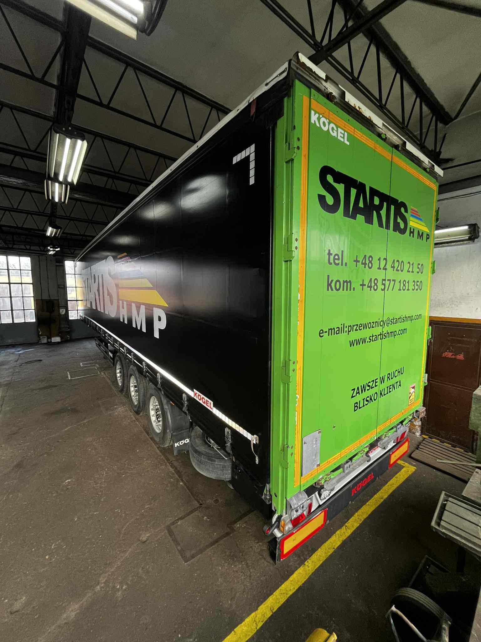 Czarna naczepa Kogel z zielonym tyłem, oklejona reklamą Startis HMP, widoczna w warsztacie. Kontrastowe kolory i wyraźne logo.