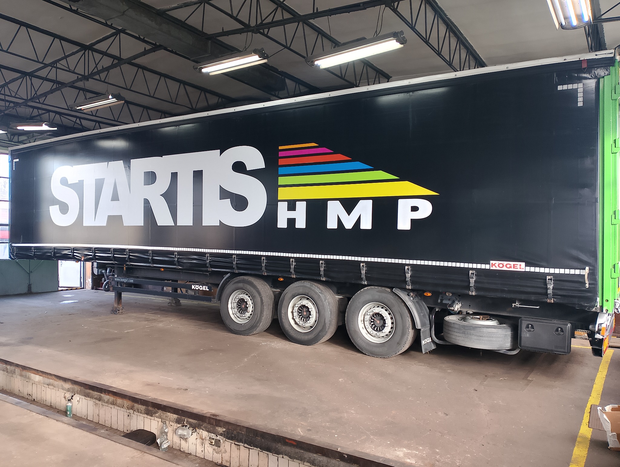 Czarna naczepa z białym napisem 'STARTIS HMP' i kolorowymi paskami, zaparkowana w warsztacie. Widoczne koła i elementy konstrukcyjne. Marka naczepy: Kogel.