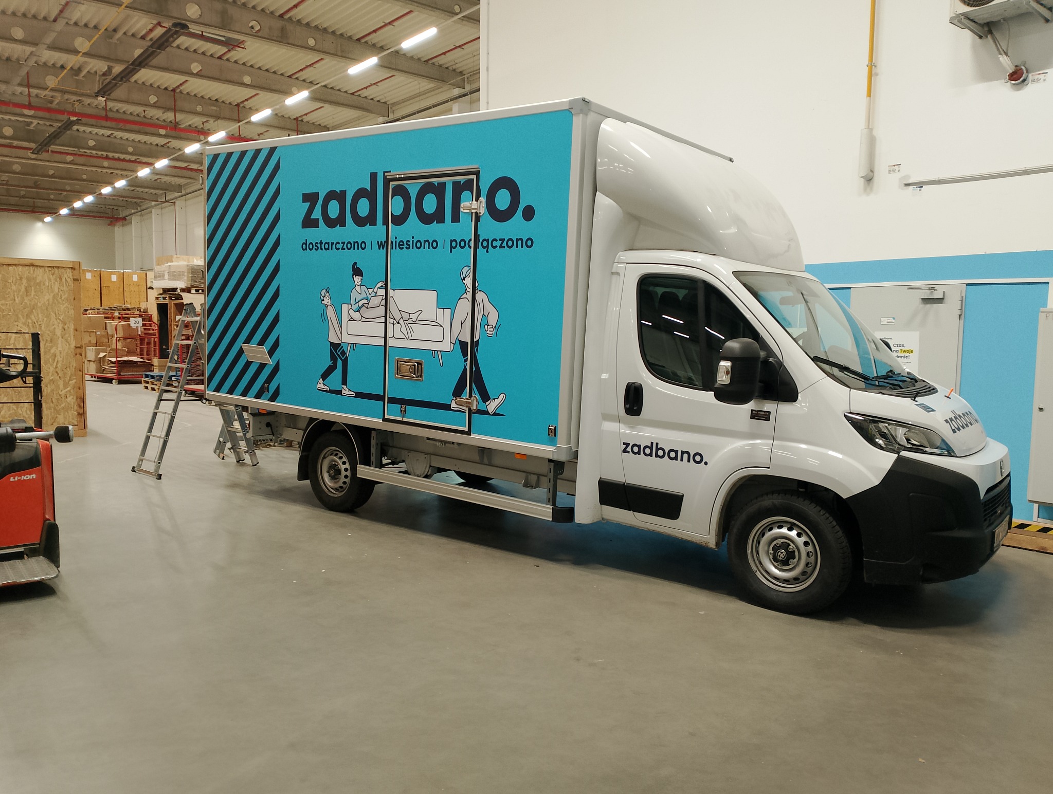 Oklejony samochód dostawczy z grafiką firmy 'zadbano. dostarczono wniesiono podłączono' na tle magazynu. Widoczna drabina i wózek widłowy.