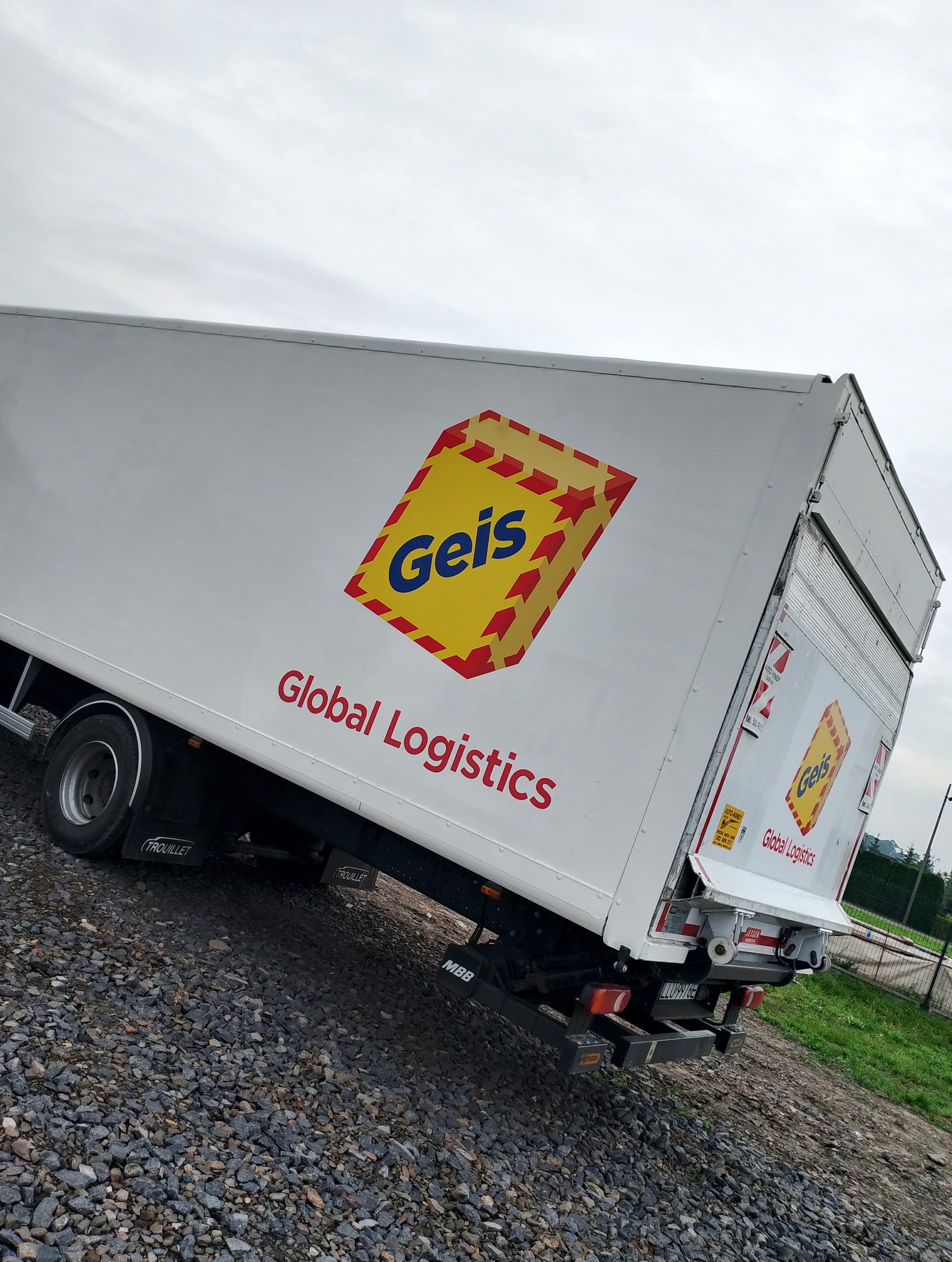 Biała ciężarówka z logo Geis Global Logistics, zaparkowana na żwirowym podłożu. Ujęcie z boku, lekko pod kątem, ukazujące tylną część pojazdu i logo firmy.