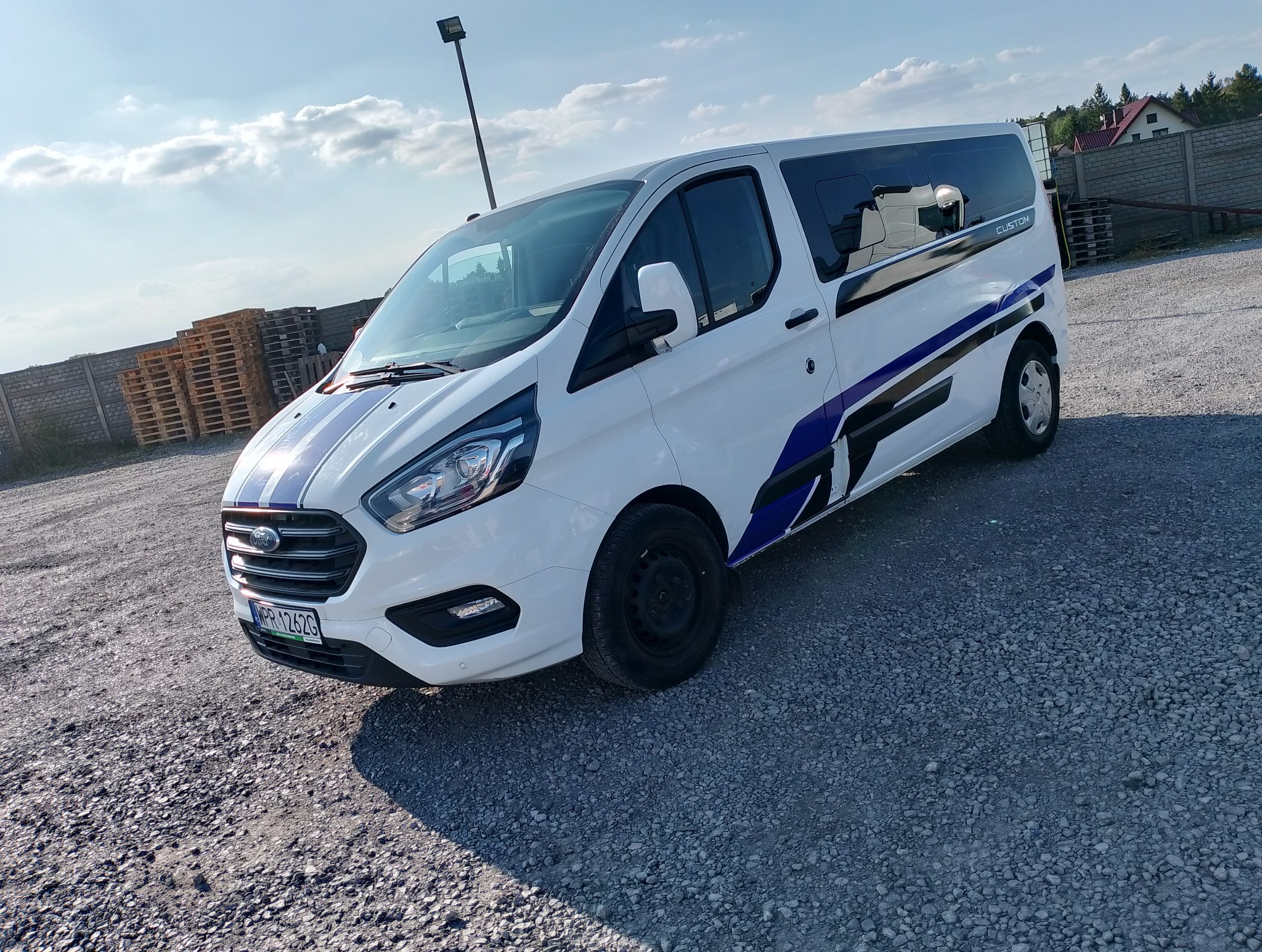 Biały Ford Transit Custom z niebiesko-czarnymi pasami na karoserii, zaparkowany na żwirowym placu. Widoczne palety w tle i błękitne niebo.