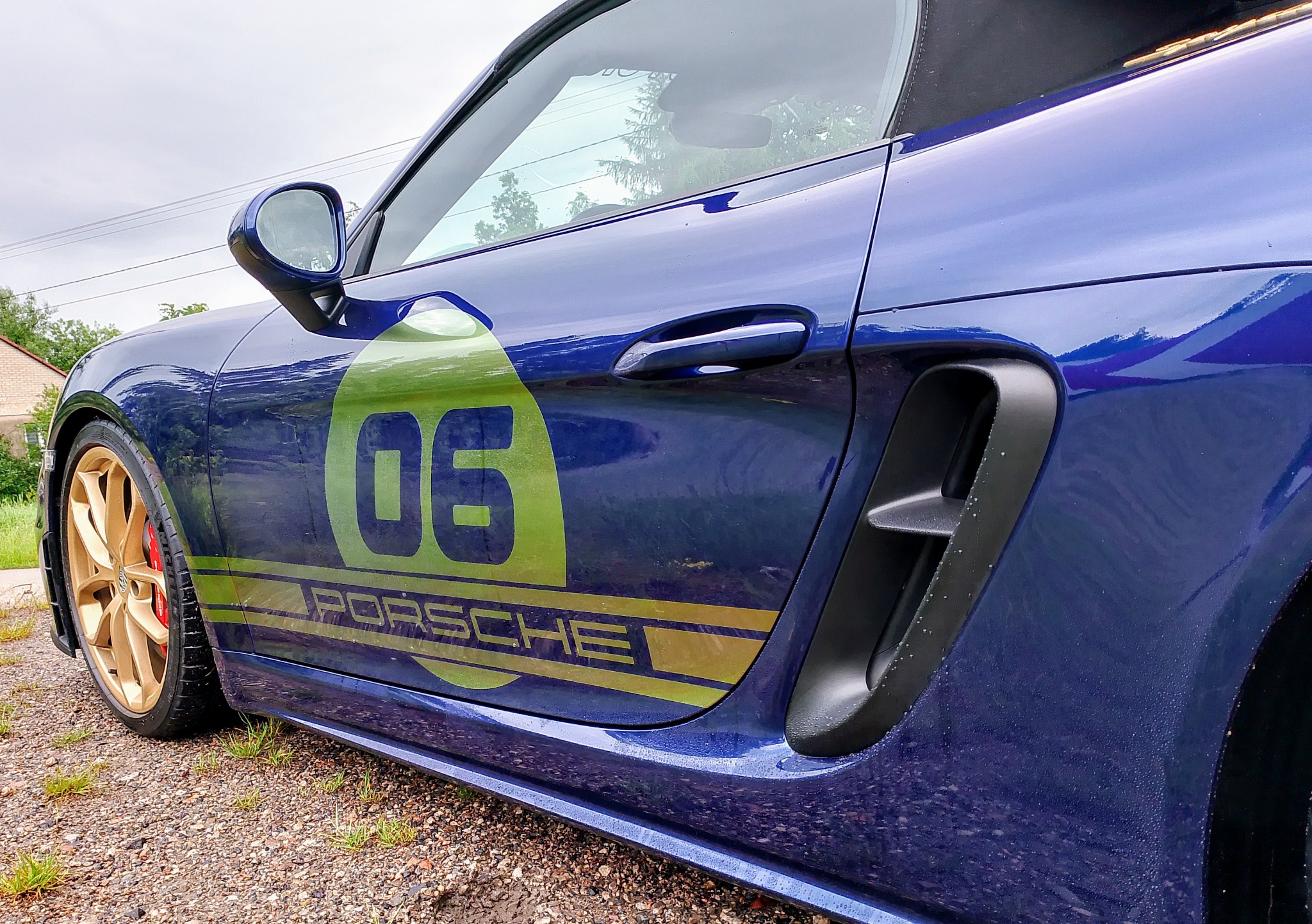 Niebieski Porsche z zielono-złotym oklejeniem bocznym: numer '06' i napis 'PORSCHE'. Złote felgi, widok z boku, mokra nawierzchnia.
