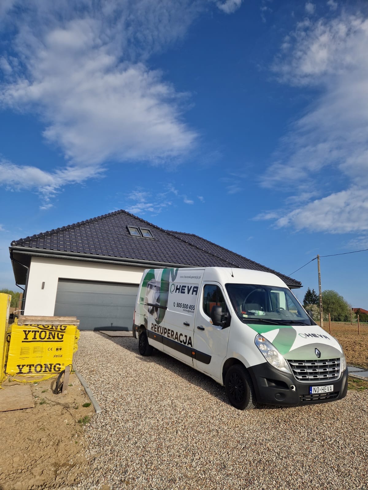 Biały van z grafiką firmy HEVA Rekuperacja na tle nowoczesnego domu z szarą bramą garażową i stertą bloczków YTONG. Błękitne niebo z chmurami w tle.
