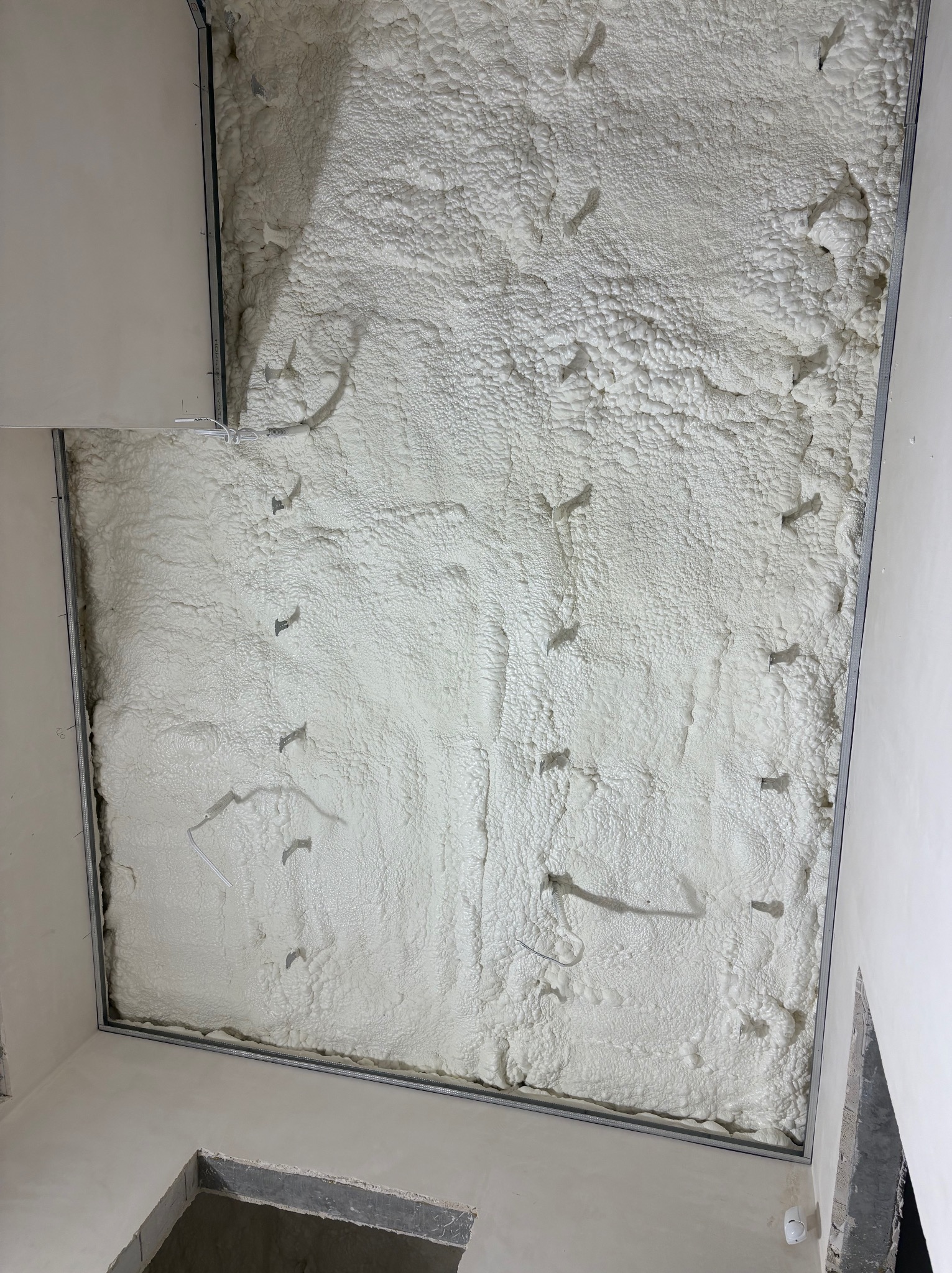 Poddasze po aplikacji białej pianki izolacyjnej, widoczne metalowe profile konstrukcyjne i wystające przewody. Nieregularna struktura pianki, surowy stan wykończenia.