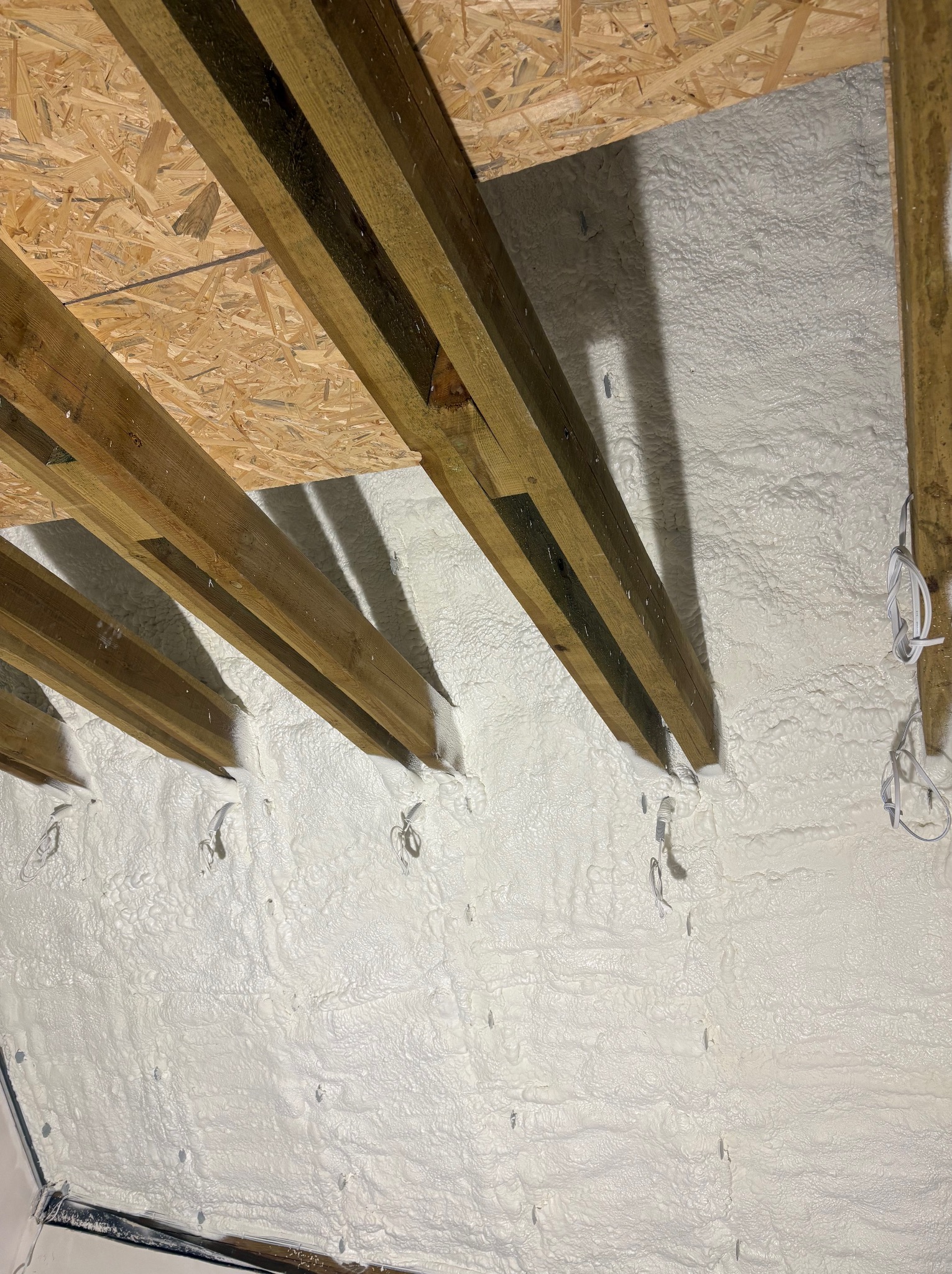 Poddasze z drewnianą konstrukcją i białą, nierówną warstwą izolacyjną, widoczne przewody. Ujęcie z dołu, podkreślające strukturę izolacji.