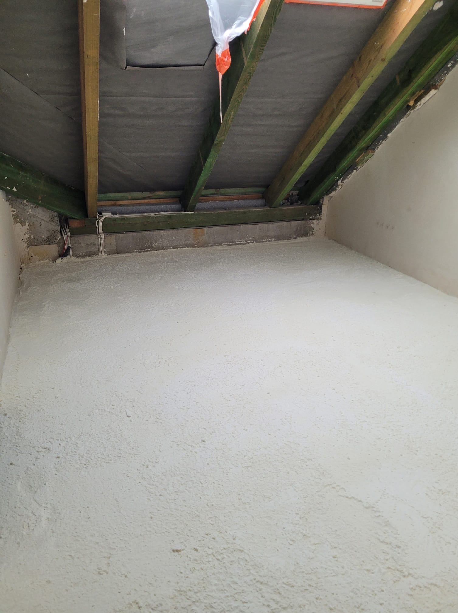 Poddasze po natrysku izolacji, widoczne drewniane elementy konstrukcyjne i szara membrana. Biała, chropowata powierzchnia izolacji pokrywa ściany i skosy.