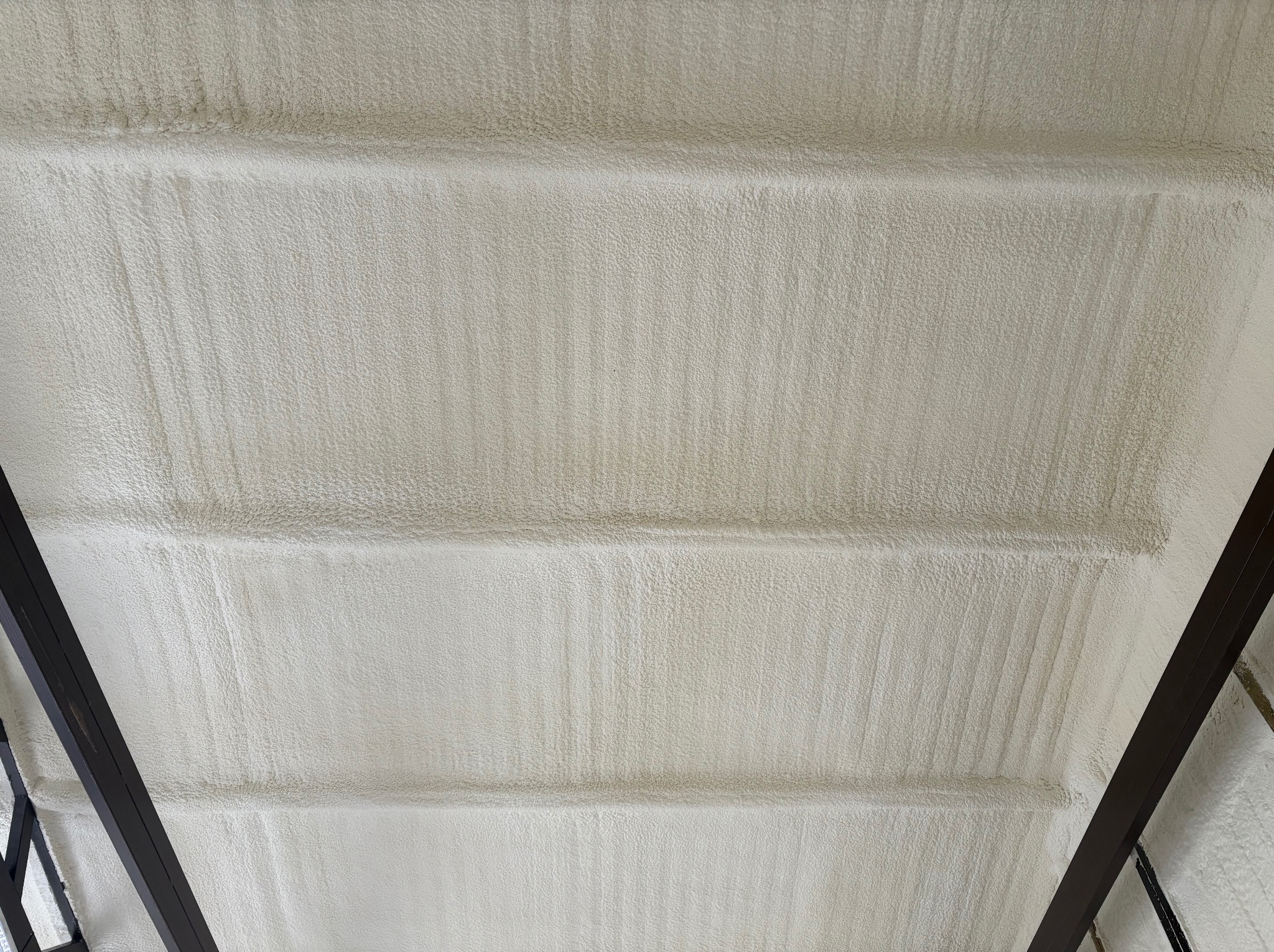 Poddasze z widoczną strukturą natryskowej izolacji, równomiernie pokrytą pianą. Elementy konstrukcji dachu w ciemnym kolorze. Tekstura piany przypomina gęstą, porowatą powierzchnię.