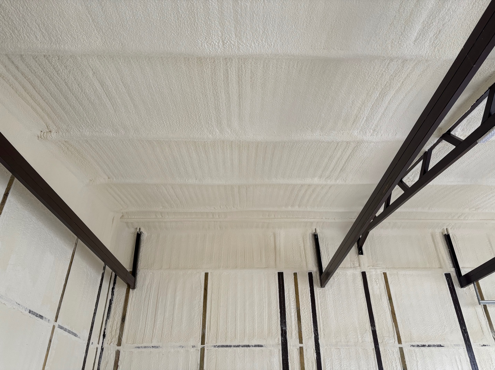 Poddasze po natrysku pianki PUR, widoczne elementy konstrukcji dachu. Biała, porowata powierzchnia izolacji, brązowe metalowe elementy mocujące.