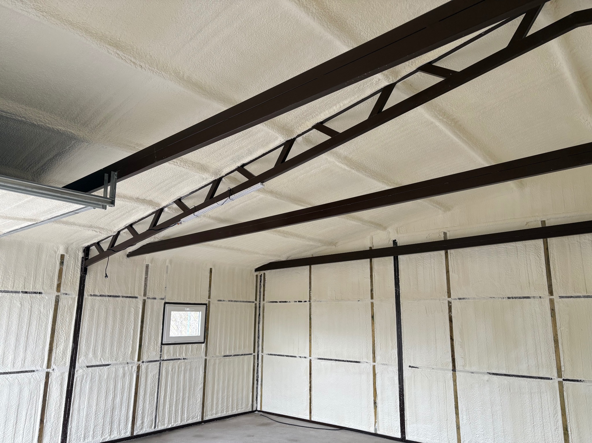 Wnętrze garażu po natrysku pianki izolacyjnej na ściany i sufit, widoczne brązowe elementy konstrukcyjne i małe okno. Biała, teksturowana powierzchnia izolacji.