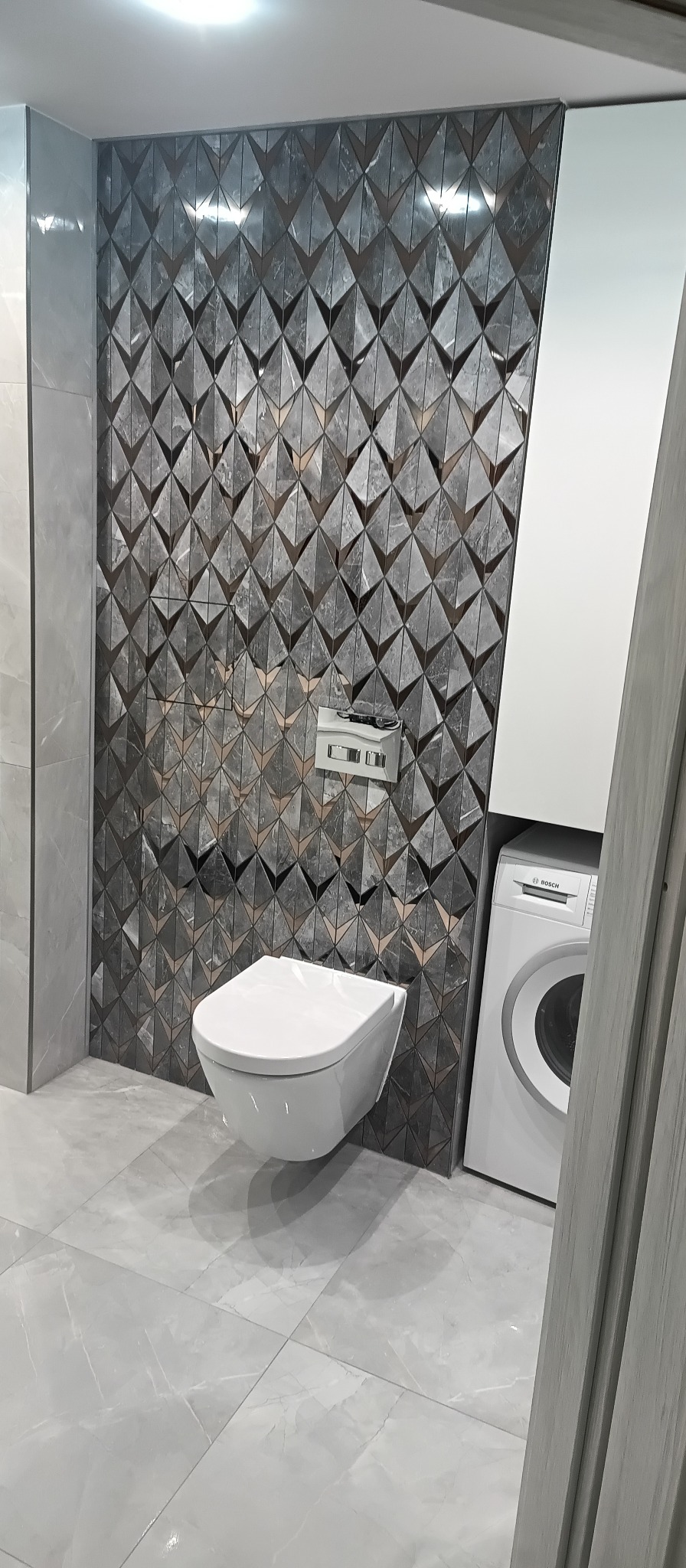 Nowoczesna łazienka z geometryczną mozaiką na ścianie, podwieszana toaleta i pralka. Szare płytki podłogowe. Minimalistyczny design.