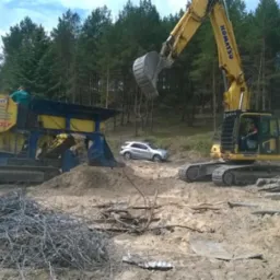 Żółta koparka Komatsu z łyżką w górze i niebiesko-żółta kruszarka do gruzu na placu budowy, w tle las i srebrny SUV.
