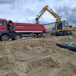 Czaj-Trans - Czerwona ciężarówka KH-Kipper i żółta koparka Liebherr 314 podczas prac ziemnych na placu budowy, z widocznym piaskiem i kamieniami na pierwszym planie.