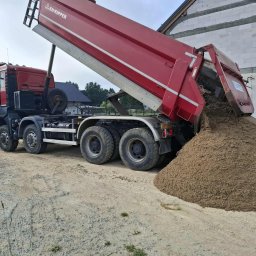 Czaj-Trans - Czerwona wywrotka z otwartą skrzynią ładunkową, wysypująca piasek na plac budowy. W tle budynek w trakcie budowy z białych bloczków.