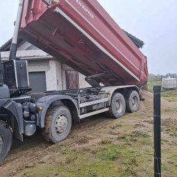 Czaj-Trans - Czerwona wywrotka KH-KIPPER wysypuje piasek na działce z budynkiem w tle. Błotniste podłoże, częściowo widoczne ogrodzenie z drutu.