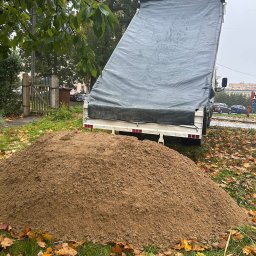Czaj-Trans - Kupa piasku na trawie z liśćmi, przyczepa z plandeką w tle, jesienna sceneria. Widok z bliska, neutralne oświetlenie.