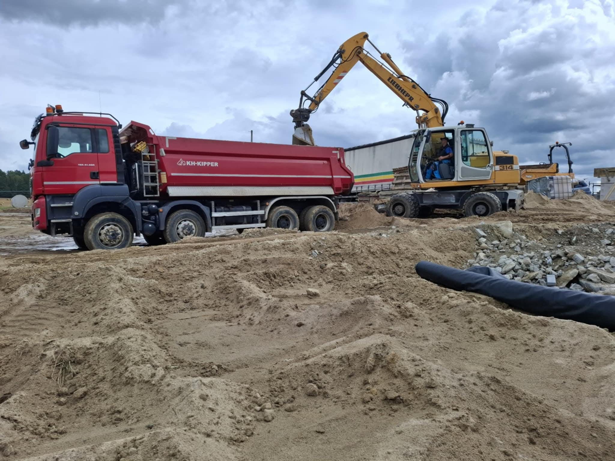 Czerwona ciężarówka KH-Kipper i żółta koparka Liebherr 314 podczas prac ziemnych na placu budowy, z widocznym piaskiem i kamieniami na pierwszym planie.