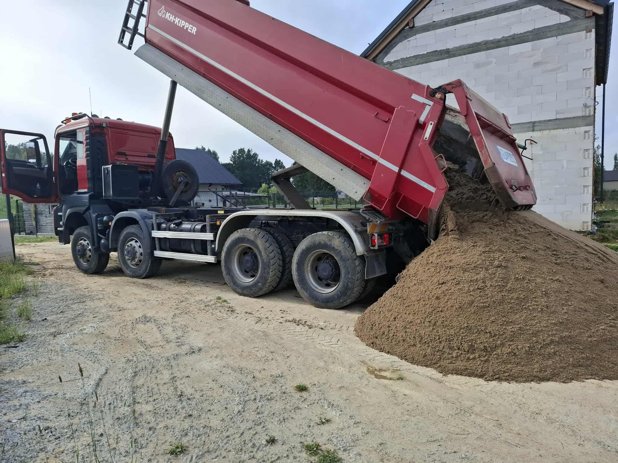 Czerwona wywrotka z otwartą skrzynią ładunkową, wysypująca piasek na plac budowy. W tle budynek w trakcie budowy z białych bloczków.
