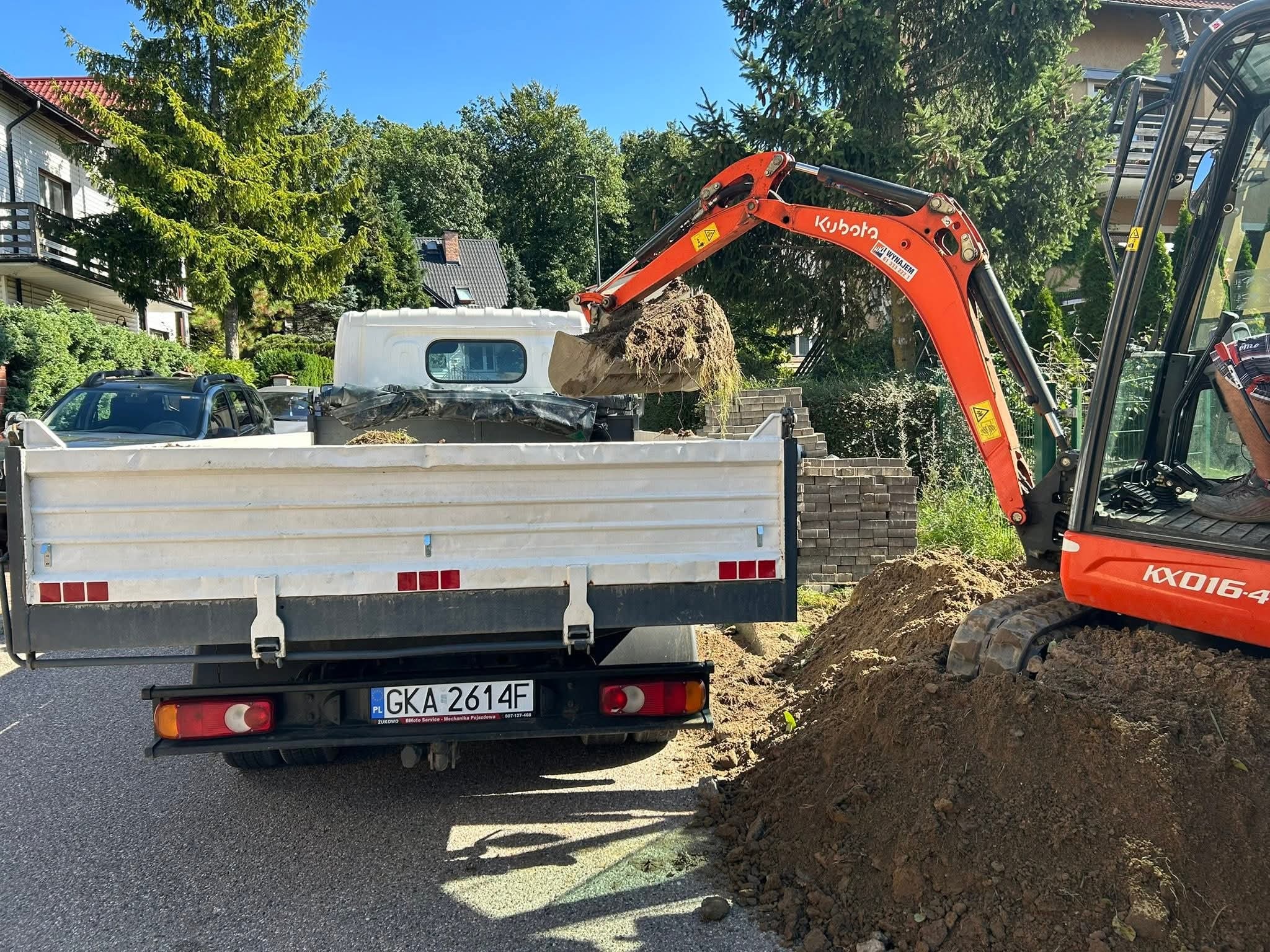 Pomarańczowa mini koparka Kubota nabiera ziemię do białej wywrotki na tle zieleni i zabudowań. Widoczna tablica rejestracyjna pojazdu.