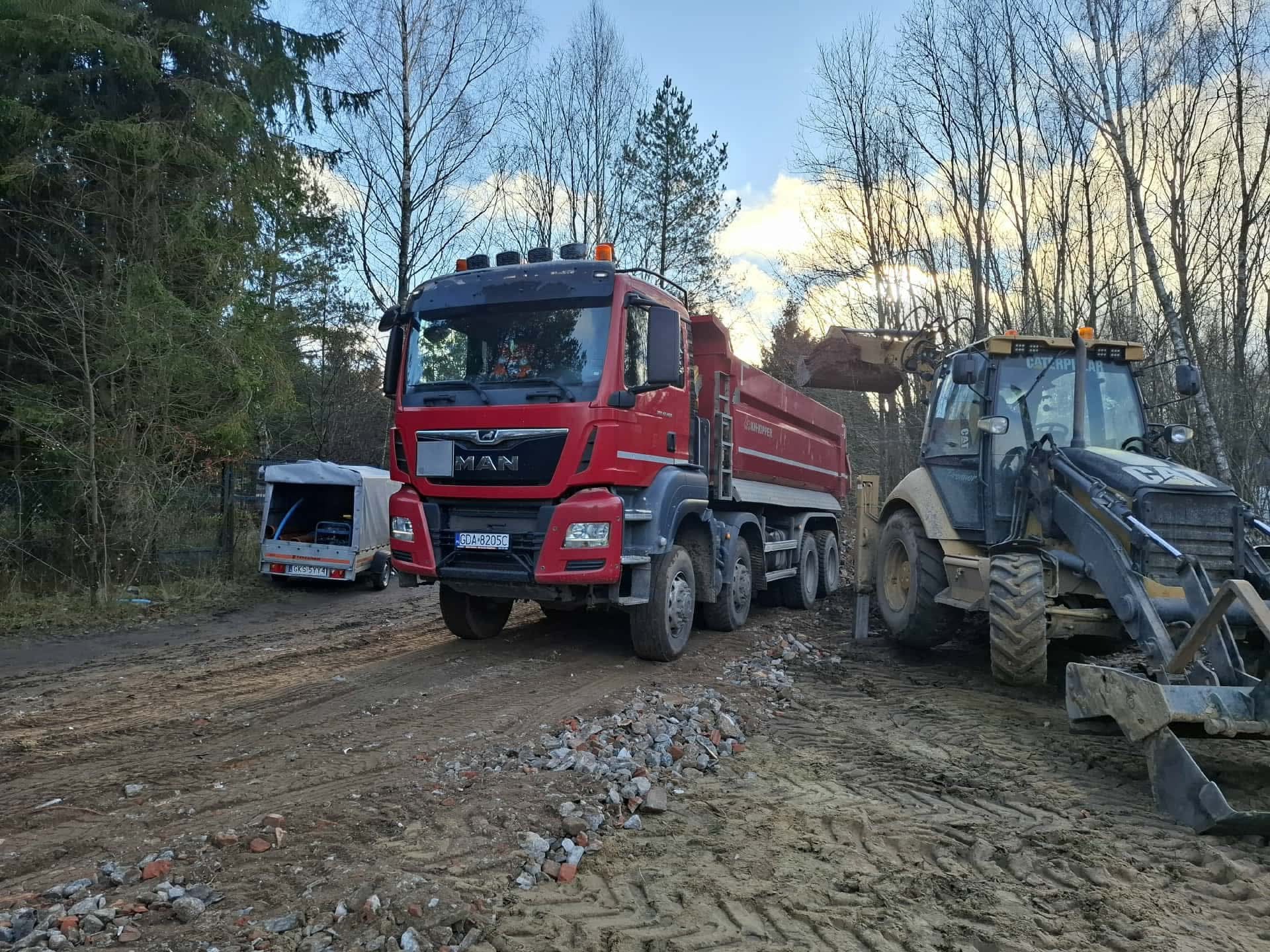 Czerwona ciężarówka MAN i koparko-ładowarka Caterpillar na placu budowy, w tle zalesiony teren i przyczepa. Widoczne ślady opon i gruz na ziemi.