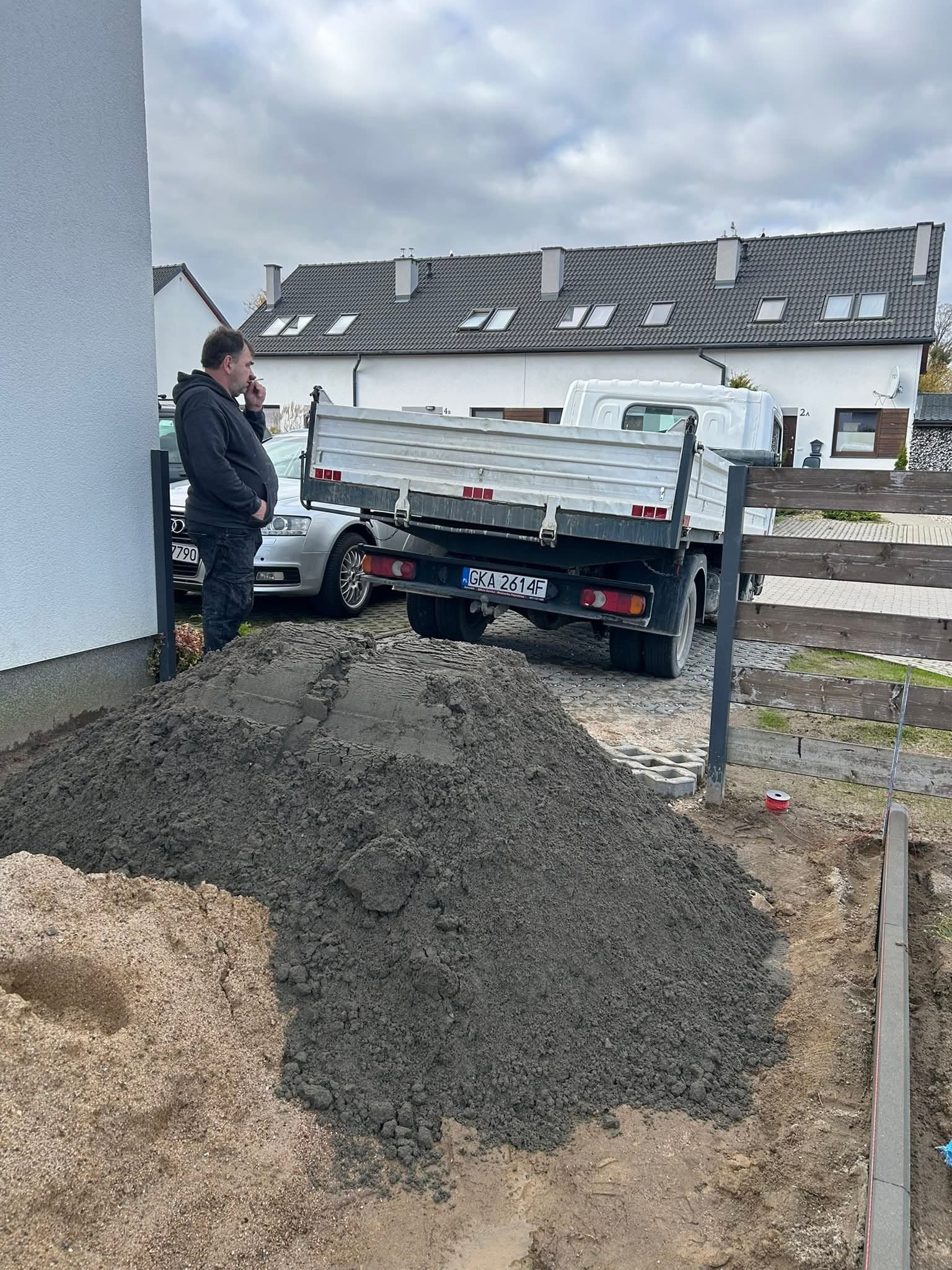 Dostawa materiałów sypkich: Góra piasku i żwiru obok ciężarówki na posesji, widoczny fragment krawężnika i pracownik w tle. Realizacja w trakcie prac ziemnych.