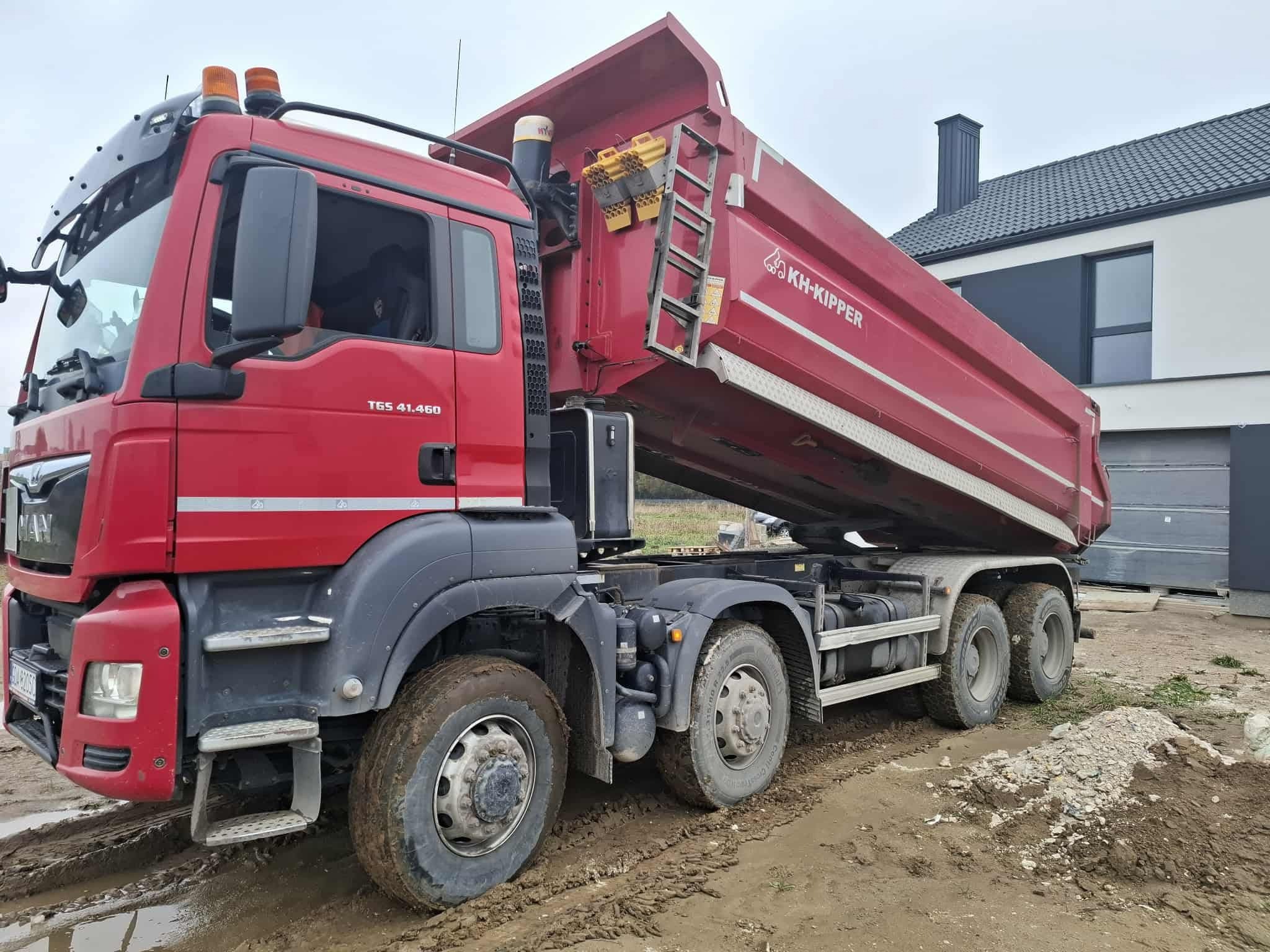 Czerwona wywrotka KH-Kipper na błotnistym terenie budowy, z podniesioną skrzynią ładunkową. W tle nowoczesny dom z ciemną dachówką i jasną elewacją.