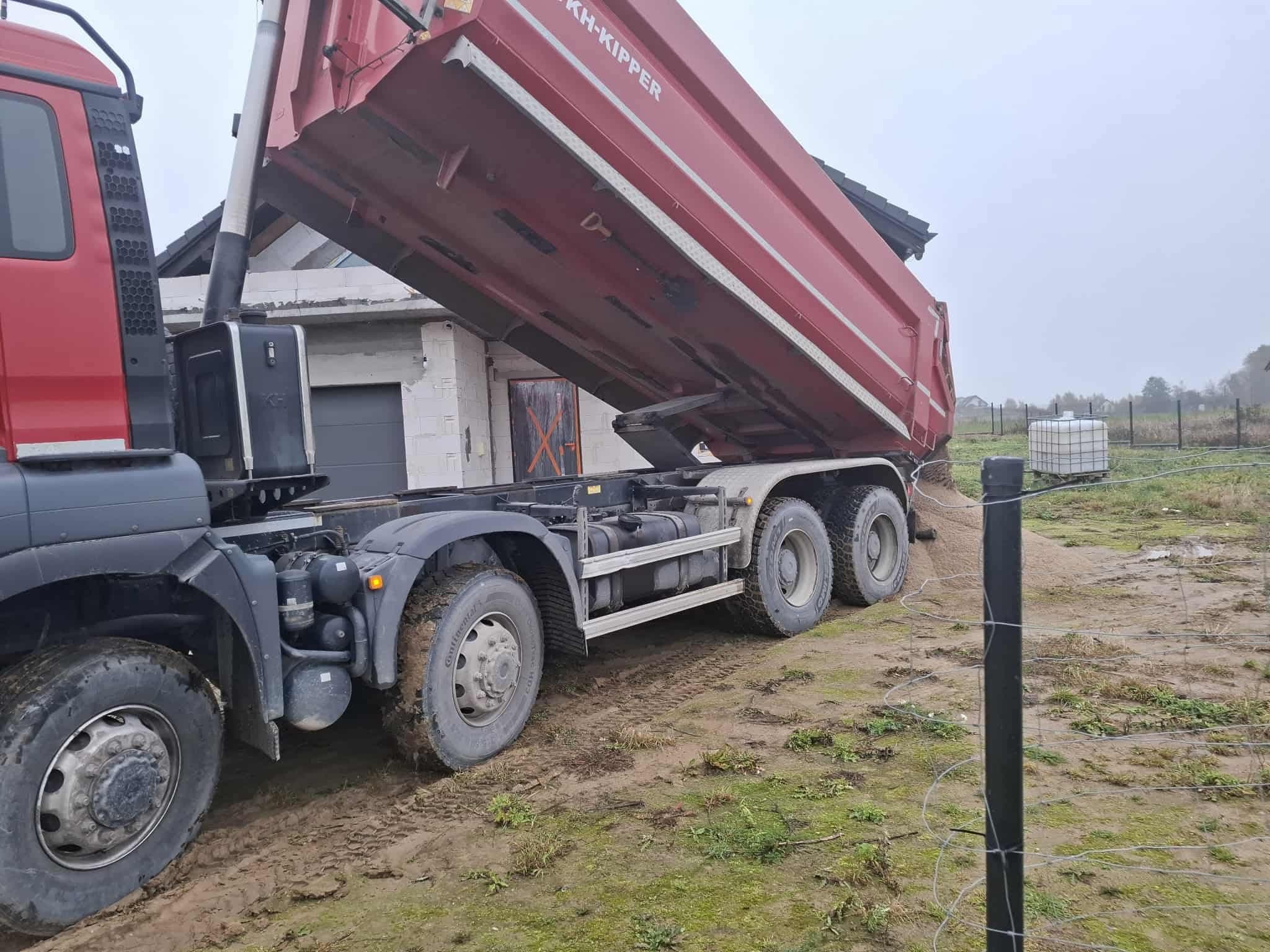 Czerwona wywrotka KH-KIPPER wysypuje piasek na działce z budynkiem w tle. Błotniste podłoże, częściowo widoczne ogrodzenie z drutu.