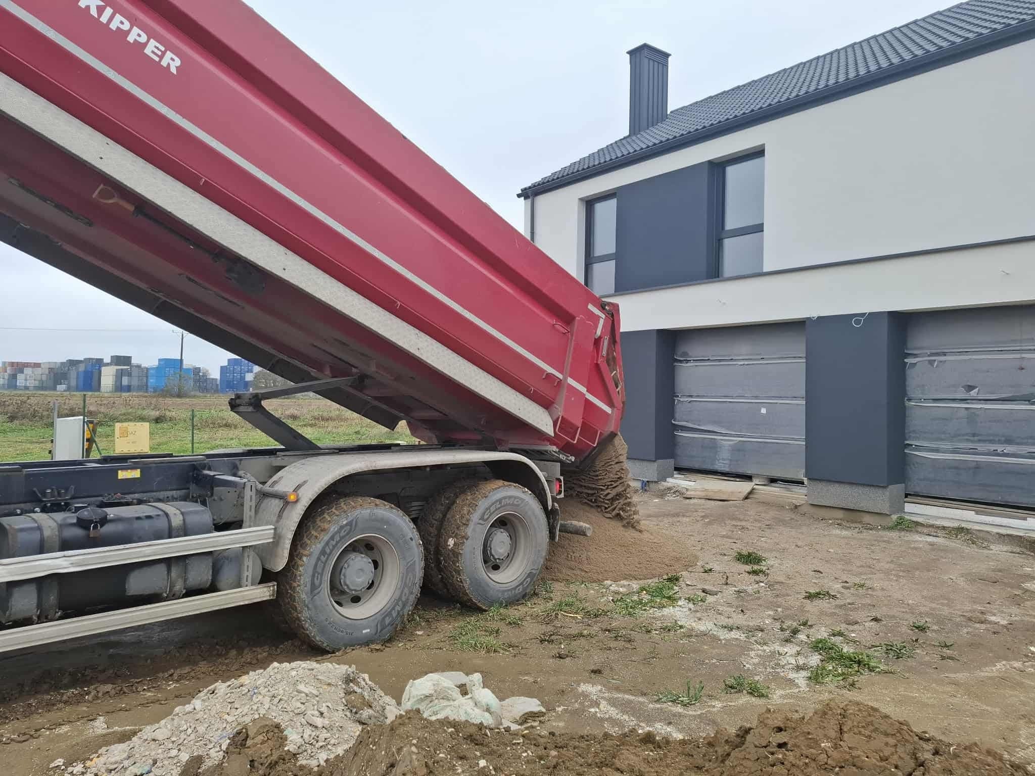 Czerwona wywrotka w trakcie wysypywania piasku na posesji przed nowoczesnym domem z szarą stolarką okienną i bramą garażową.