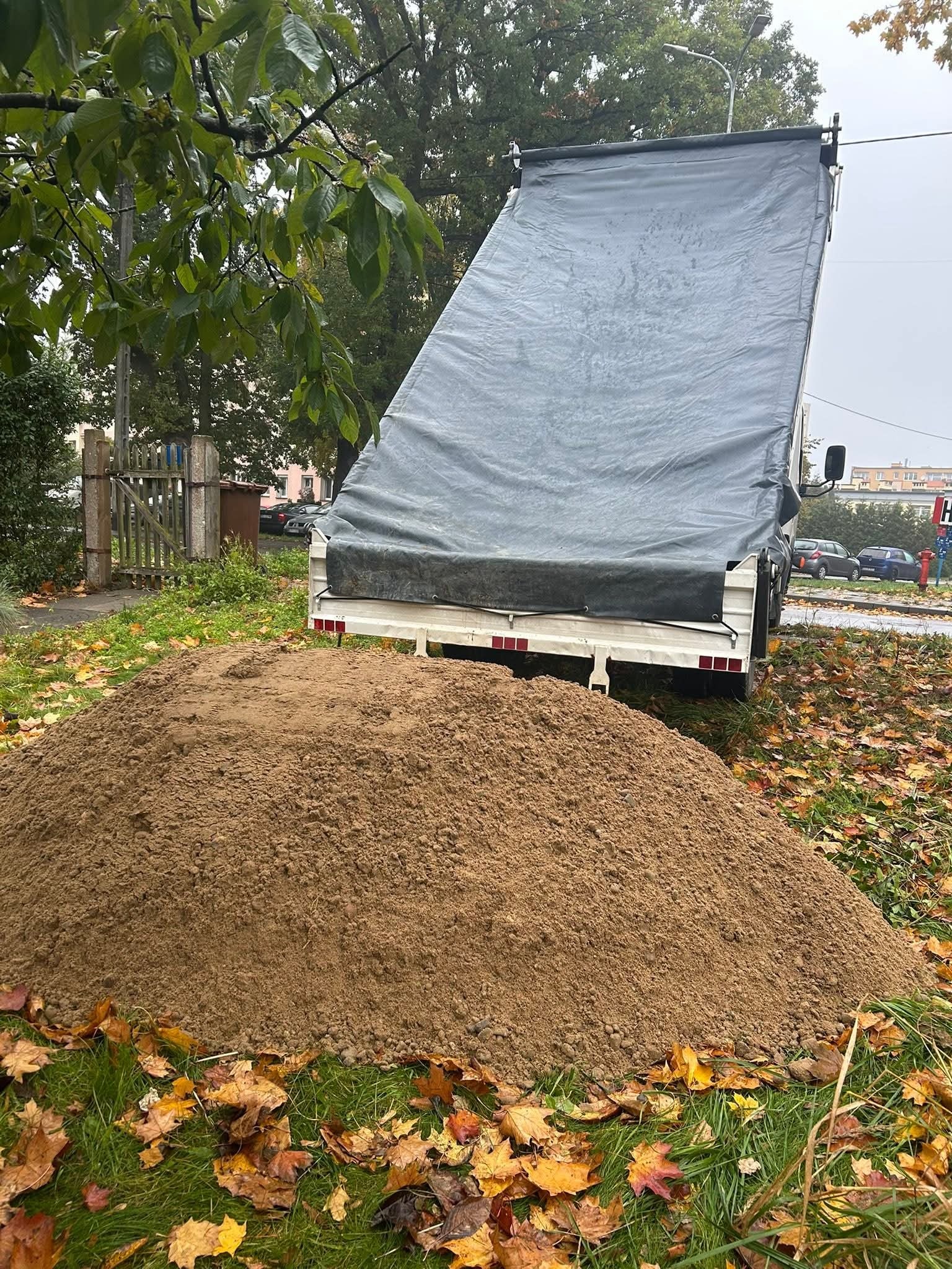 Kupa piasku na trawie z liśćmi, przyczepa z plandeką w tle, jesienna sceneria. Widok z bliska, neutralne oświetlenie.