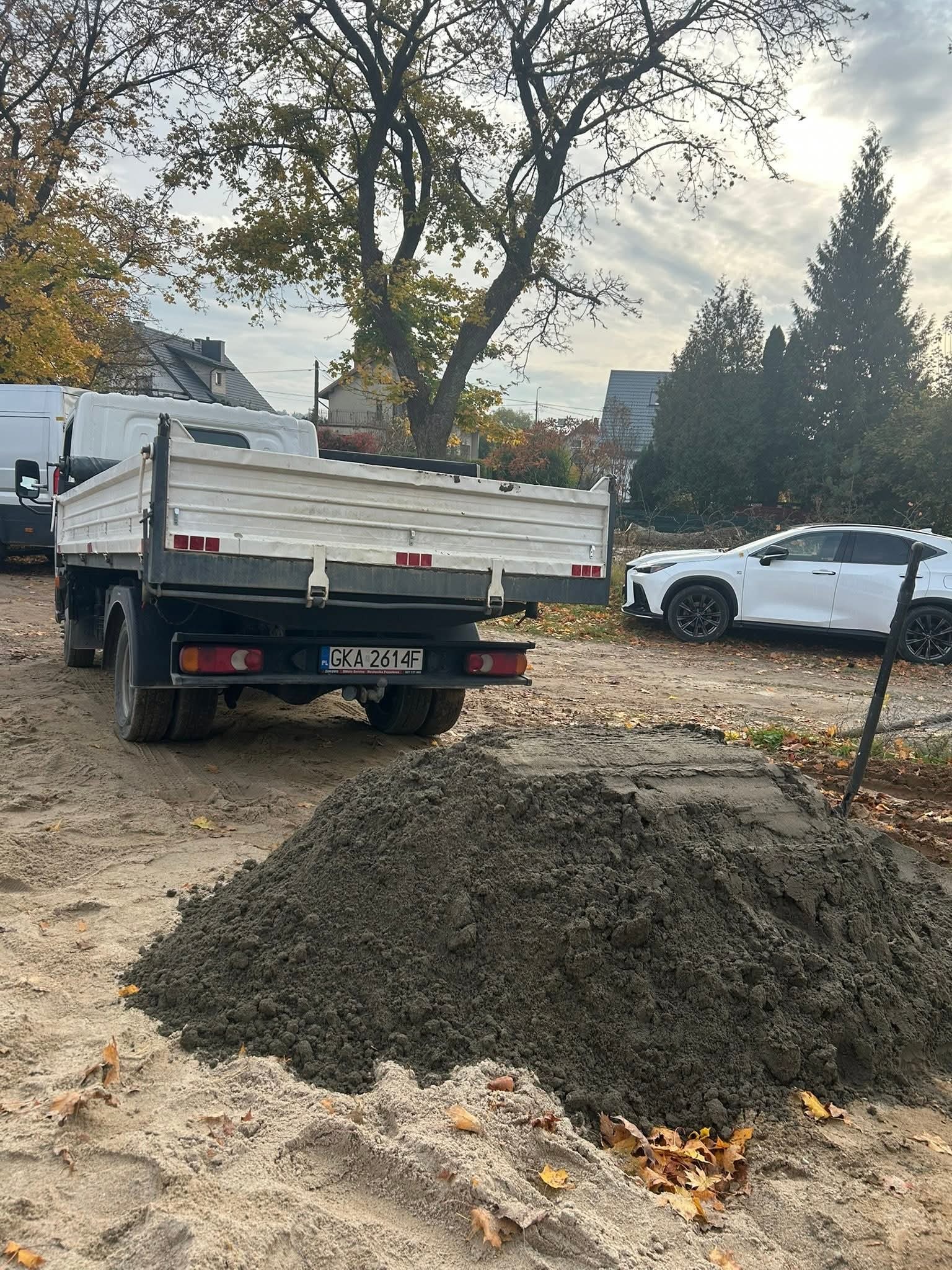 Ciężarówka z wywrotką i sterta szarego materiału na ziemi, prawdopodobnie piasku lub żwiru, na tle domu i białego samochodu osobowego.