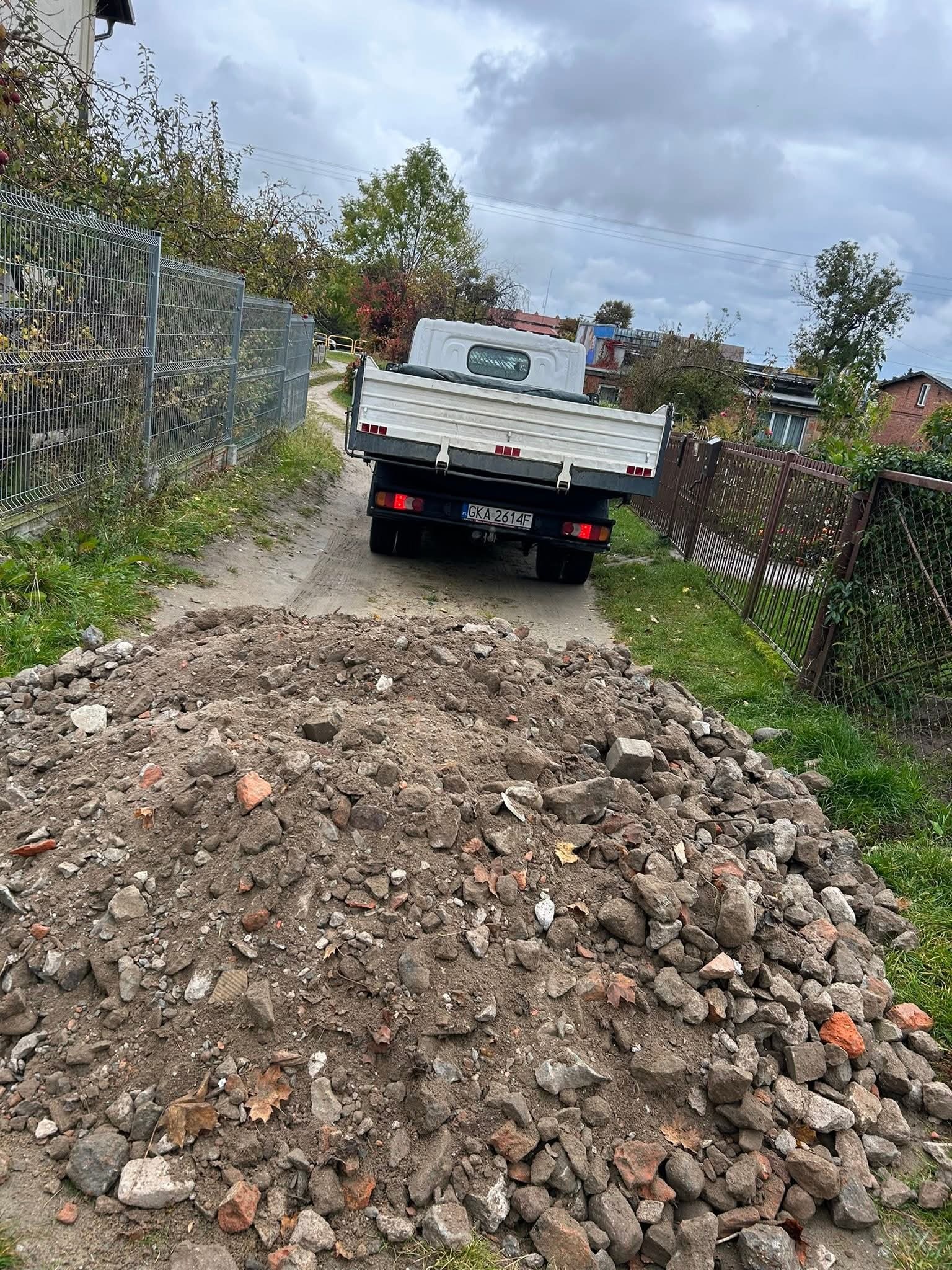 Kiprująca wywrotka na gruntowej drodze, za stertą gruzu i kamieni, w tle ogrodzenie posesji i zabudowania. Widok z tyłu pojazdu.