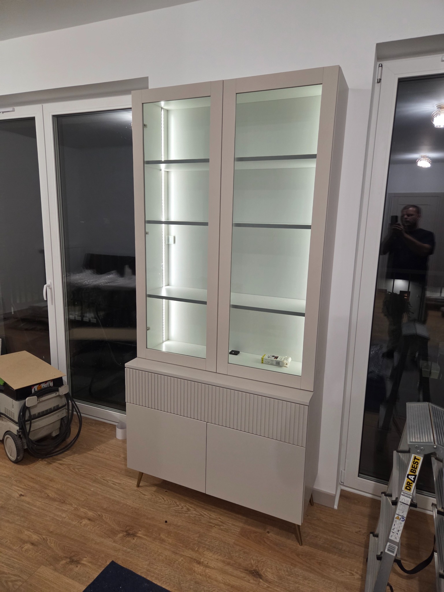 Nowoczesna witryna z podświetlanymi półkami, beżowe fronty z frezowanymi zdobieniami, ustawiona na drewnianej podłodze, widoczne okna i drabina.