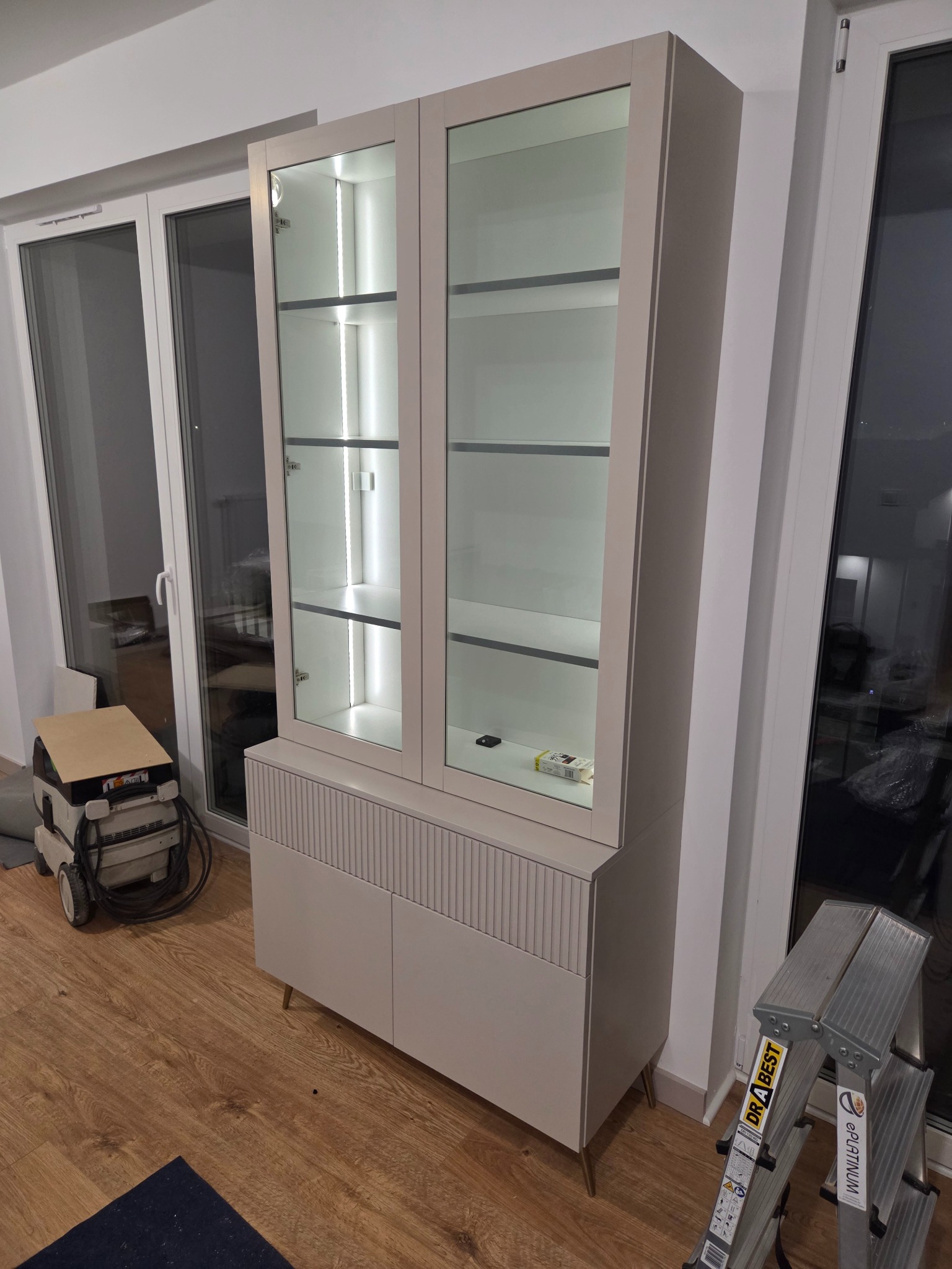 Biała witryna z przeszkleniami i oświetleniem LED, dolna część z ryflowanymi drzwiczkami na złotych nóżkach. Obok okno i drabina.