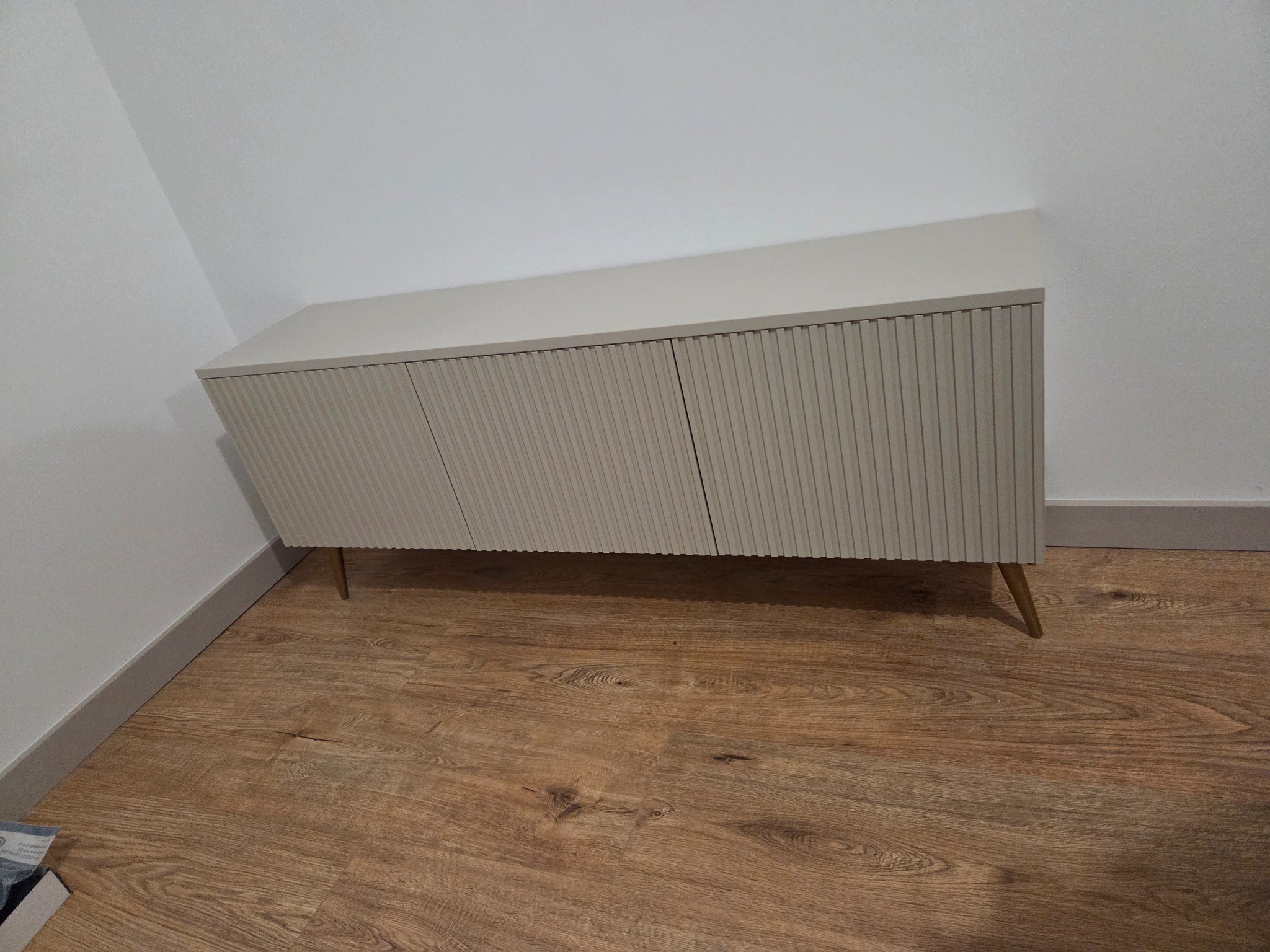 Beżowa komoda z ryflowanymi frontami, na złotych nóżkach, ustawiona na drewnianej podłodze przy białej ścianie. Minimalistyczny design.