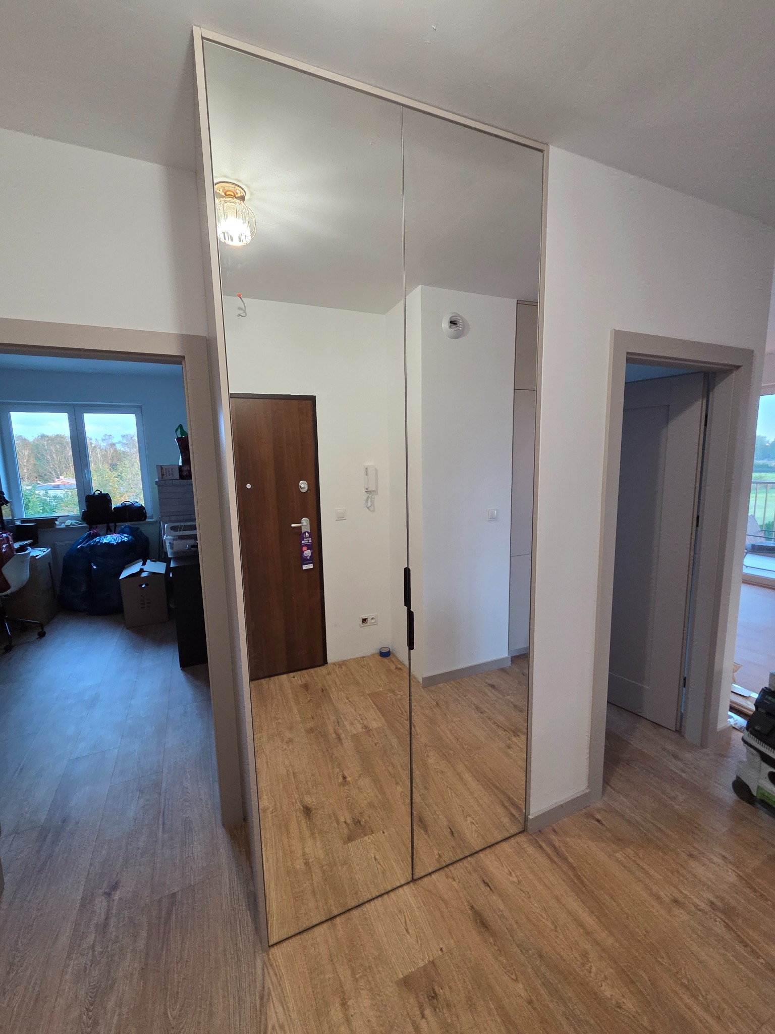 Nowoczesna szafa wnękowa z lustrzanymi drzwiami przesuwnymi, odbijająca wnętrze mieszkania. Widoczny parkiet i drzwi wejściowe w odbiciu.