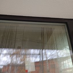 PIEŃO - BUD Damian Piechowiak - Zapocone okno z widocznymi smugami i naklejoną kartką, odbijające budynek i drzewa. Widoczny fragment sufitu i ramy okiennej.