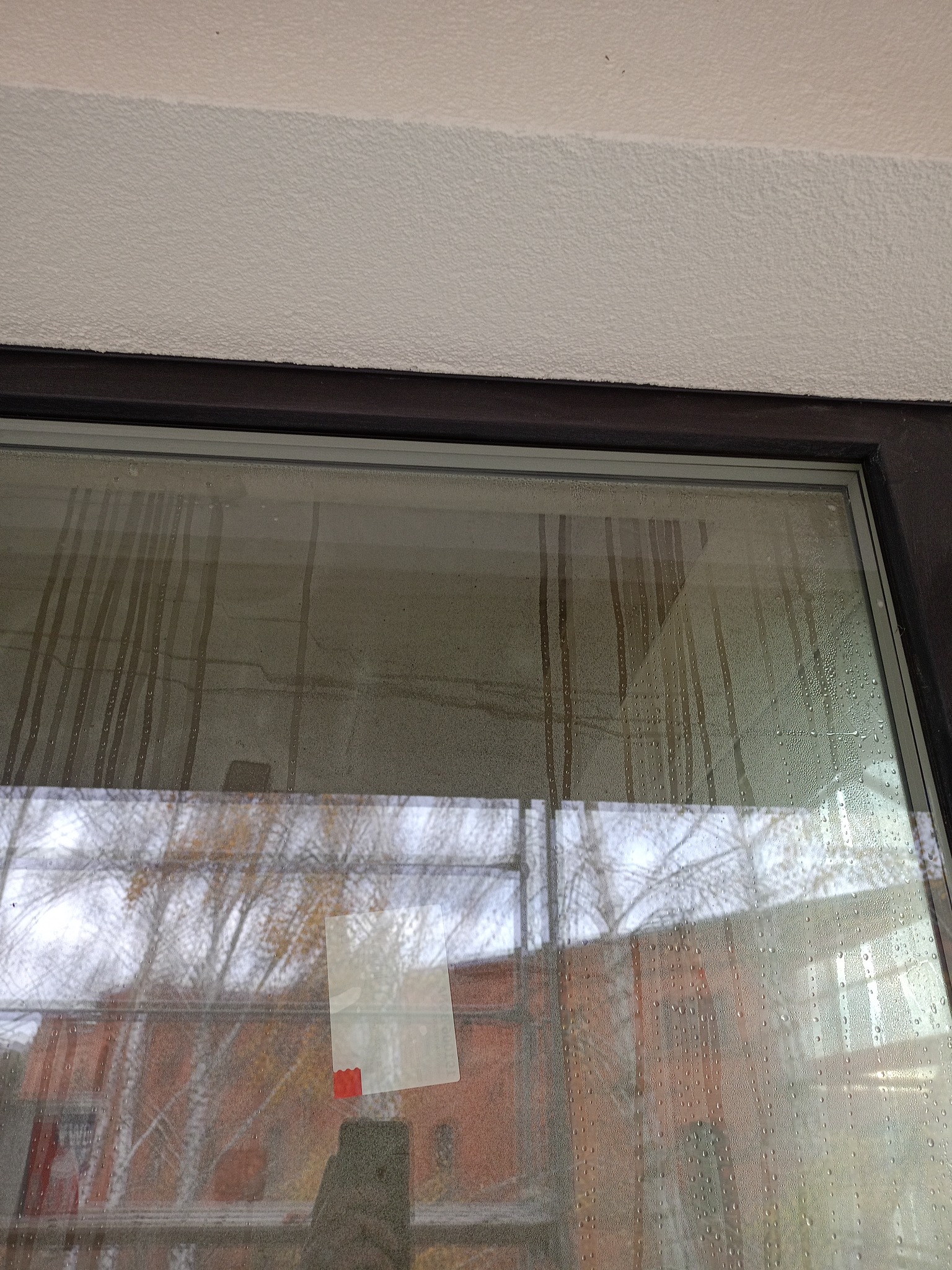 Zapocone okno z widocznymi smugami i naklejoną kartką, odbijające budynek i drzewa. Widoczny fragment sufitu i ramy okiennej.