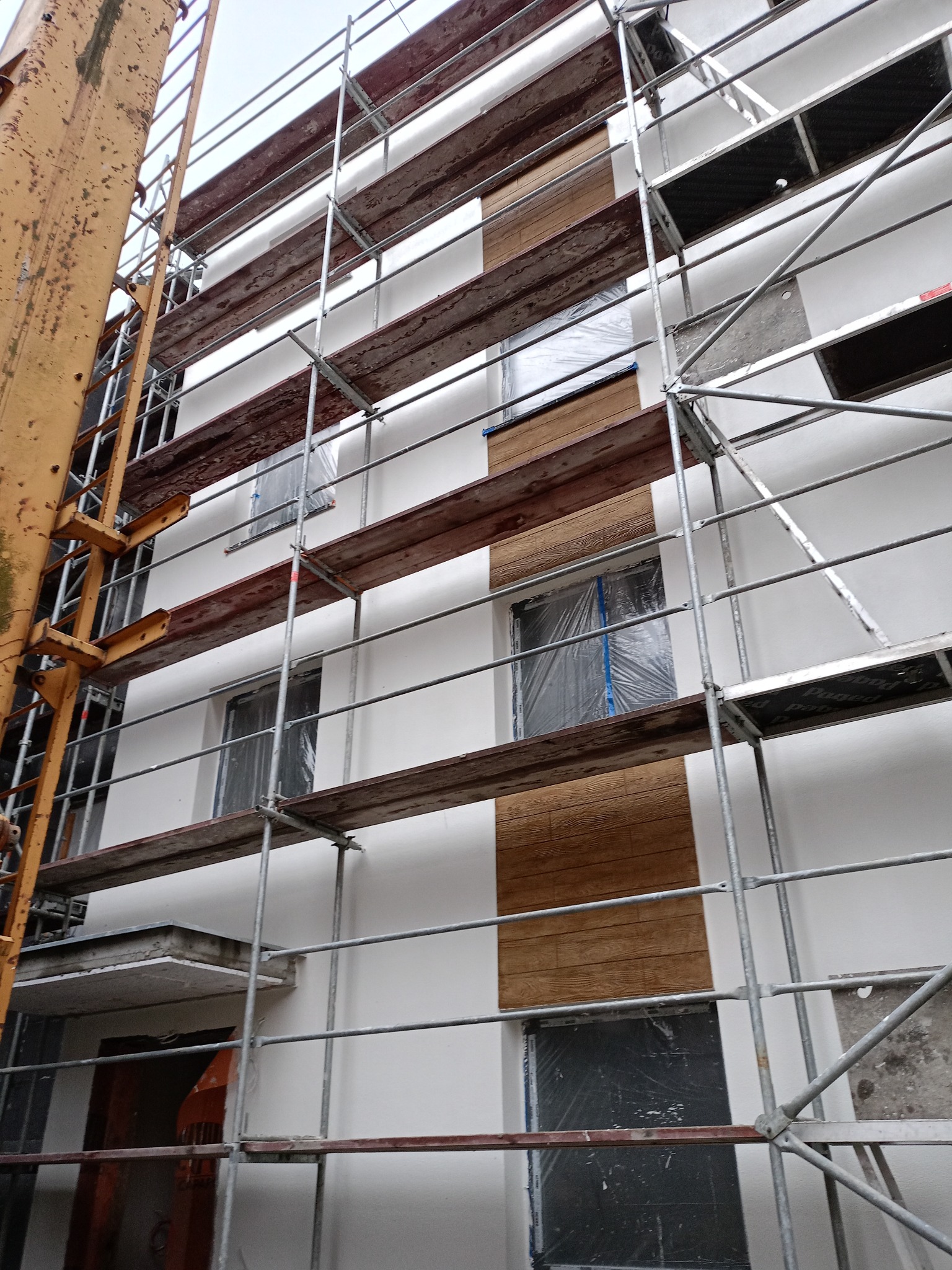 Elewacja budynku w trakcie prac wykończeniowych, z widocznymi rusztowaniami i zabezpieczonymi oknami folią. Fragment ściany z dekoracyjnym panelem imitującym drewno.