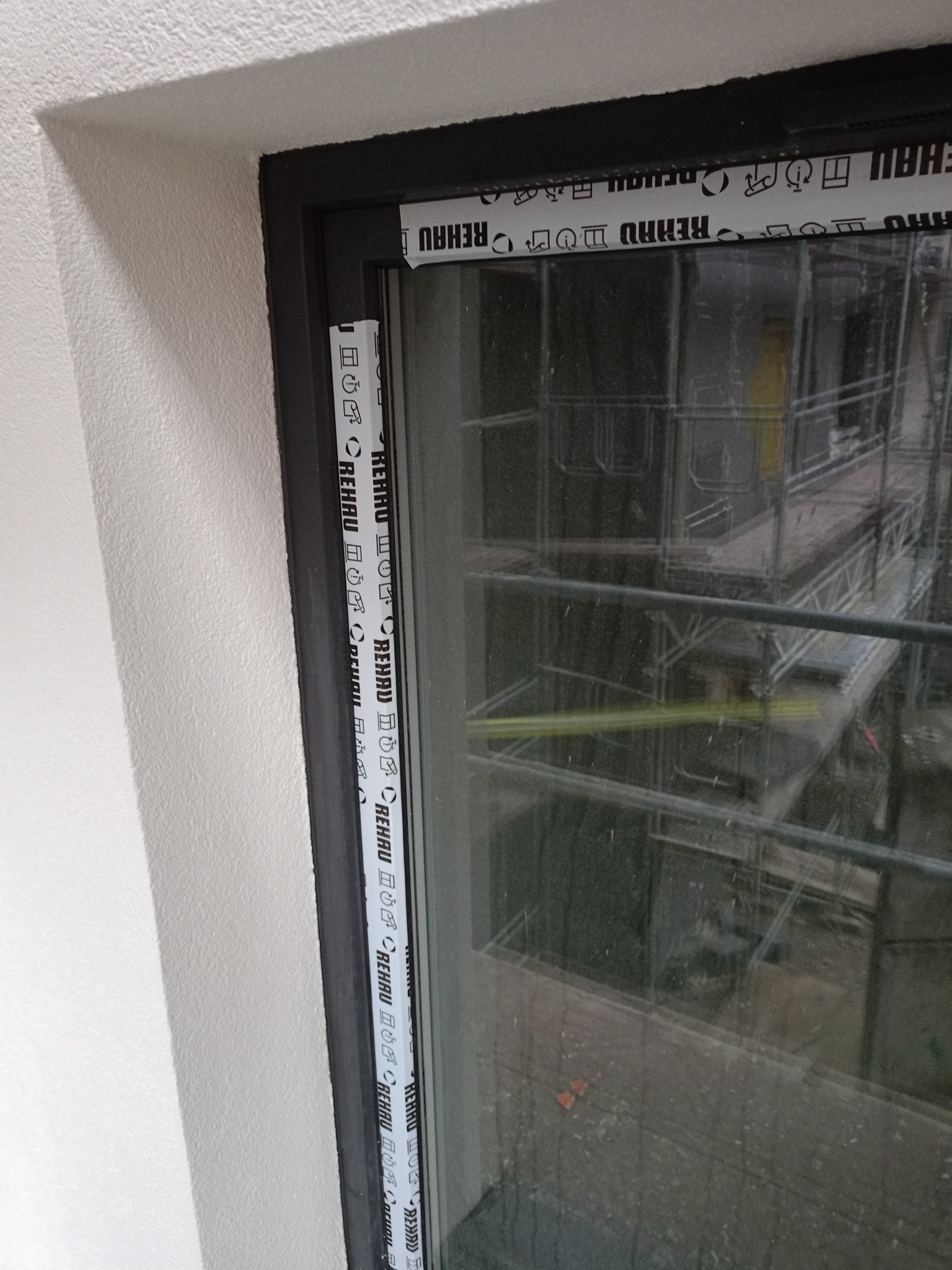 Zamontowane okno Rehau z czarną ramą w otynkowanej ścianie. Na zewnątrz widoczne rusztowania. Detal wykończenia w nowym budownictwie.