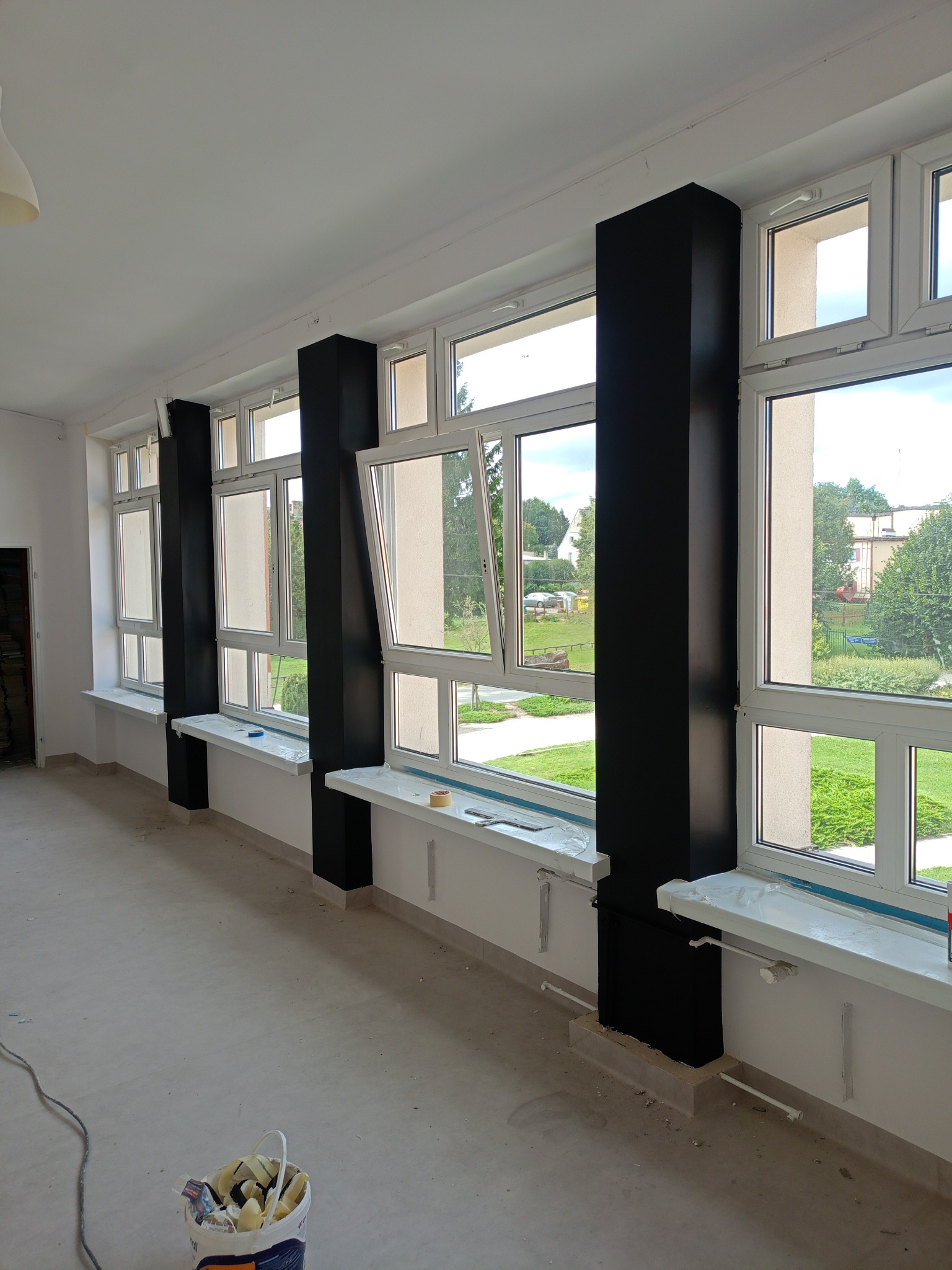 Wnętrze w trakcie prac: białe ściany, okna z czarnymi filarami, zabezpieczone folią parapety i wiadro z akcesoriami malarskimi na podłodze. Widok na zewnątrz przez otwarte okno.