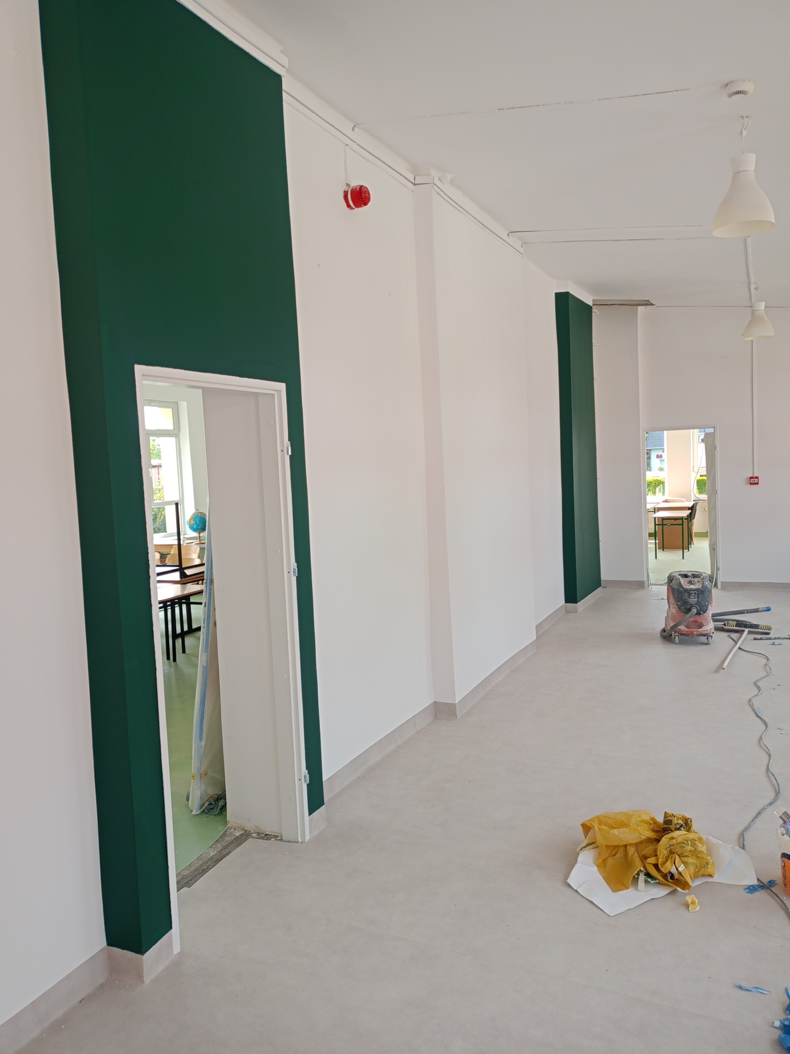 Białe ściany z zielonymi akcentami wokół drzwi w odnowionym wnętrzu. Na podłodze widoczne ślady prac malarskich i narzędzia, co sugeruje etap wykończeniowy.