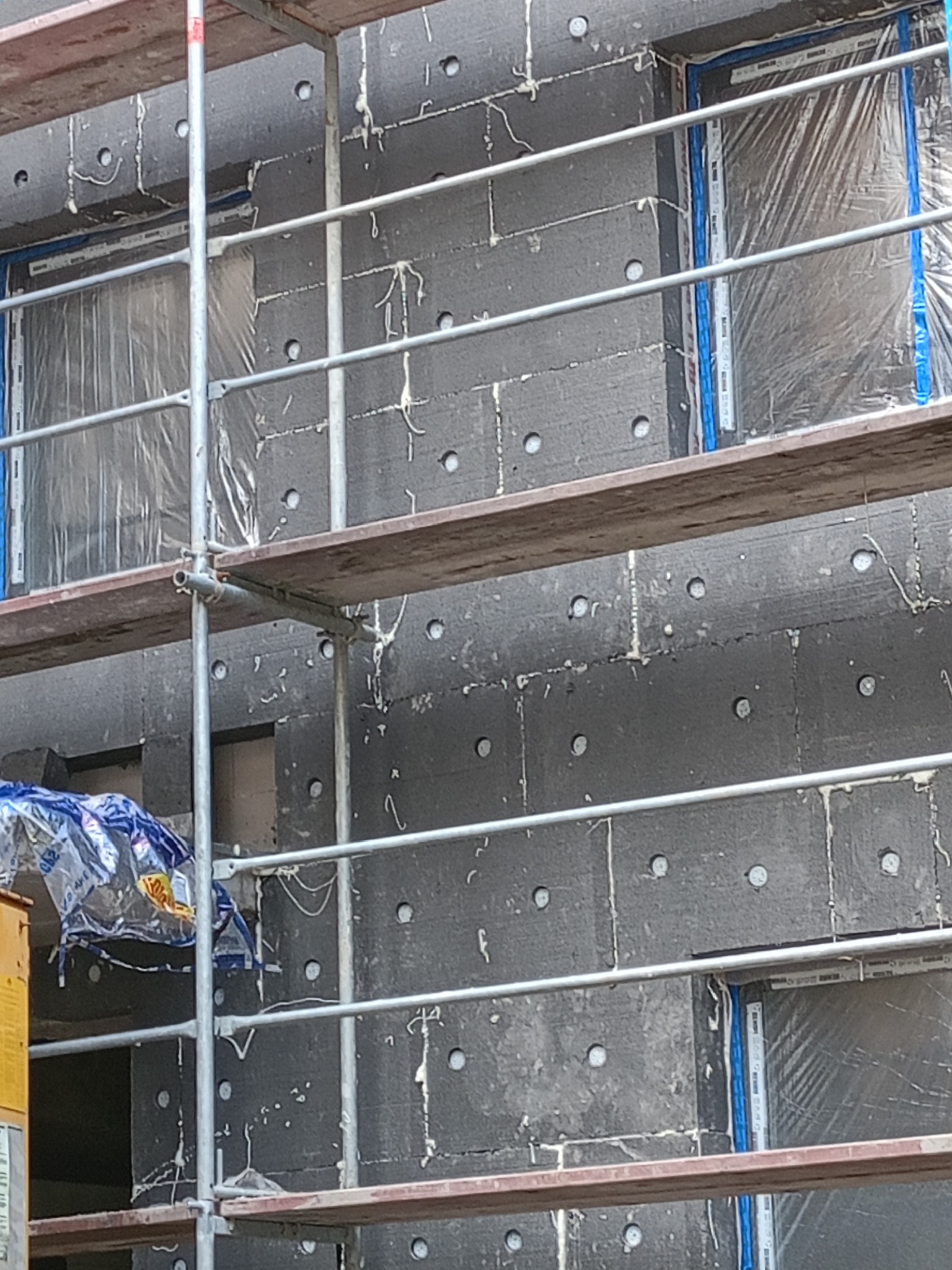 Ocieplanie grafitowym styropianem na elewacji budynku w Grudziądzu, widoczne rusztowanie i zabezpieczone okna folią. Prace w toku, widok z bliska.