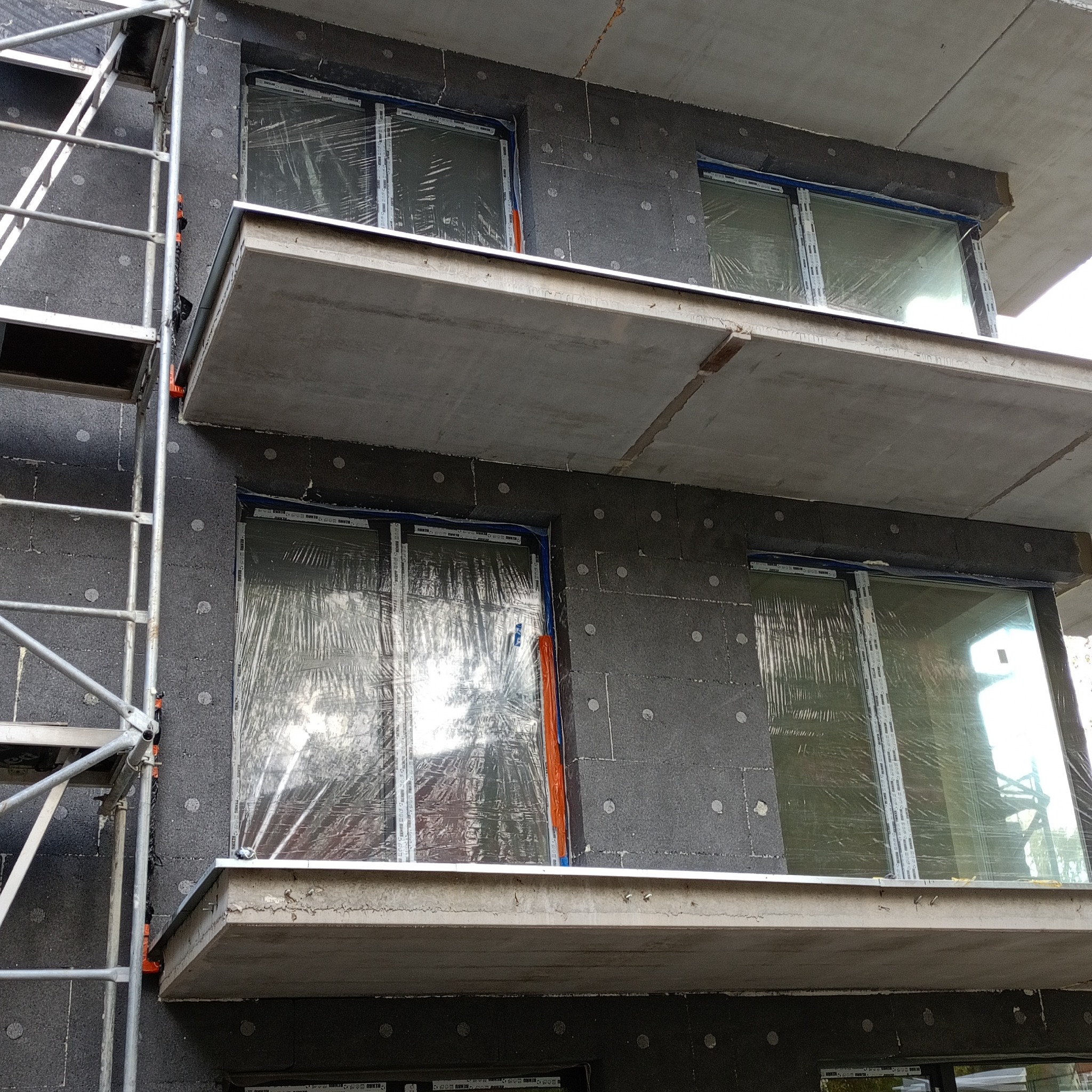 Ocieplanie budynku styropianem grafitowym, widoczne rusztowanie i okna zabezpieczone folią. Etap prac przed nałożeniem tynku.