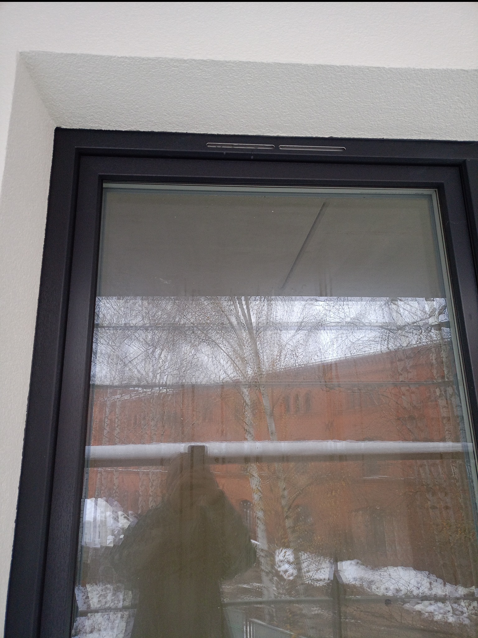 Ciemna rama okienna na tle białej ściany. W szybie odbija się budynek z czerwonej cegły i drzewa. Widoczny fragment balkonu z resztkami śniegu.