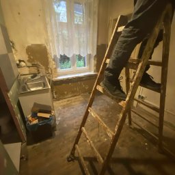 Tadeusz De - Remont starej kuchni: drabina, odsłonięte ściany, okno z firanką. Widoczny fragment zlewu i śmieci. Przygotowania do prac wykończeniowych w zaniedbanym wnętrzu.