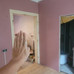 Tadeusz De - Różowa ściana z dwoma otworami drzwiowymi w trakcie wykończenia. W tle widoczna łazienka i podłoga z panelami. Ręka z pierścionkiem w kadrze.