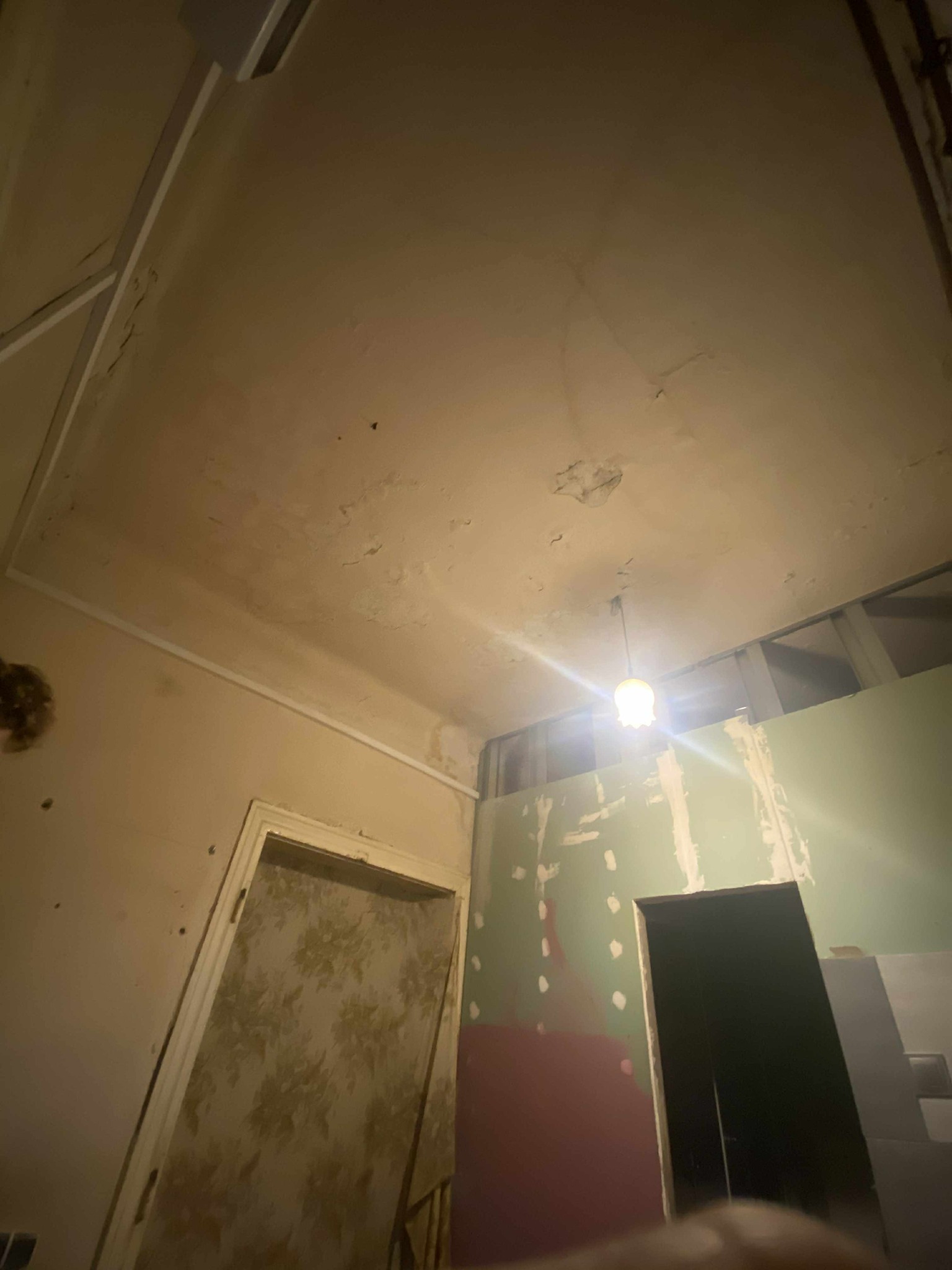 Wnętrze w trakcie remontu: widoczny sufit z plamami, ściany z fragmentami tapet i płyt karton-gips, oraz futryna drzwi. Ujęcie z dołu, oświetlenie punktowe.