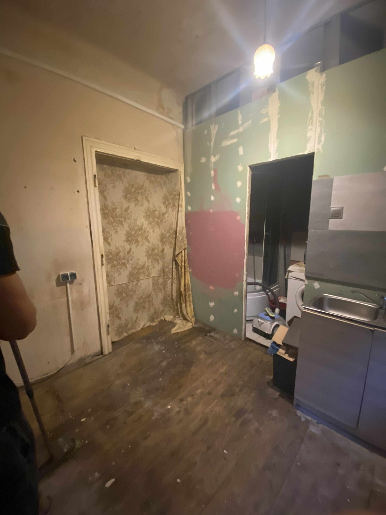 Wnętrze w trakcie remontu: stara podłoga, ściany z fragmentami tapet i śladami gładzi, widoczne instalacje. Częściowo wyposażona kuchnia w tle.