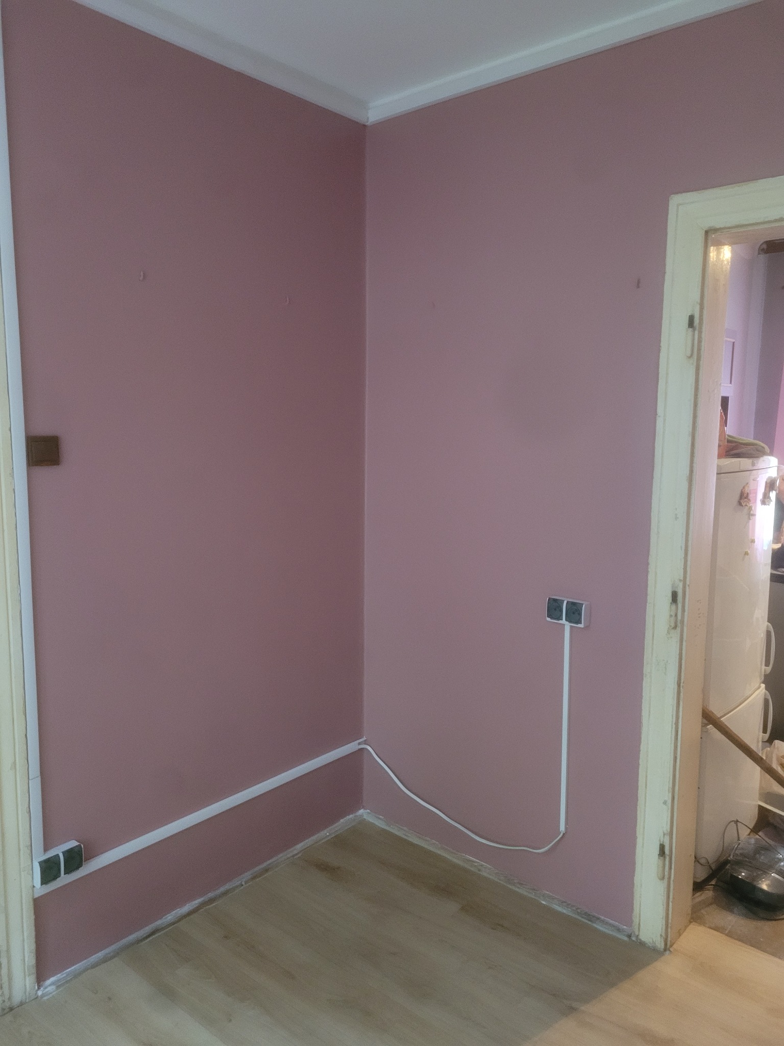 Różowe ściany po malowaniu w rogu pokoju z widocznymi gniazdkami i listwami przypodłogowymi. Podłoga z paneli. Widok na sąsiednie pomieszczenie przez otwarte drzwi.